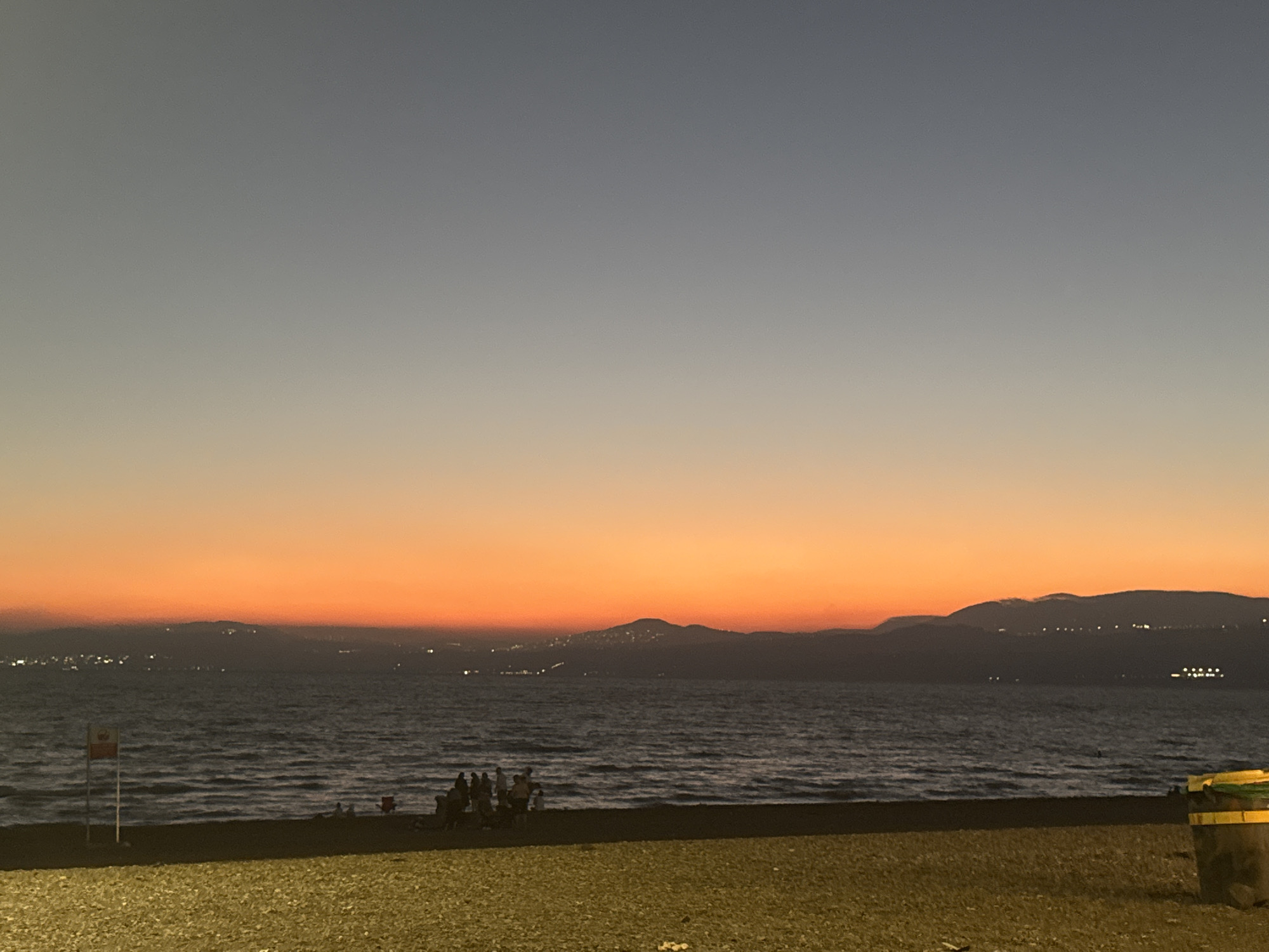 The image captures a tranquil dusk scene along a coastline, possibly in the Golan Regional Council, Israel. The sky transitions from a pale gray at the zenith to a vibrant orange hue near the horizon, indicating the setting or rising sun. A calm body of water, likely the sea, occupies the midground, with gentle waves lapping against the shore. The shoreline itself is a mix of dark sand and scattered pebbles, with a section of paved or gravel area in the foreground.
Several individuals are visible on the beach, appearing to be a small group of people gathered together. One person is seated on a red chair. A flag with a red and white emblem is planted in the sand near the water's edge. In the background, a mountainous silhouette is discernible, with scattered lights suggesting a distant settlement or city. The water itself shows a few faint reflections of lights from the distant shore. To the far right of the image, a large, dark waste bin with a yellow lid is partially visible. The overall atmosphere is serene and peaceful, characteristic of a quiet evening by the sea.