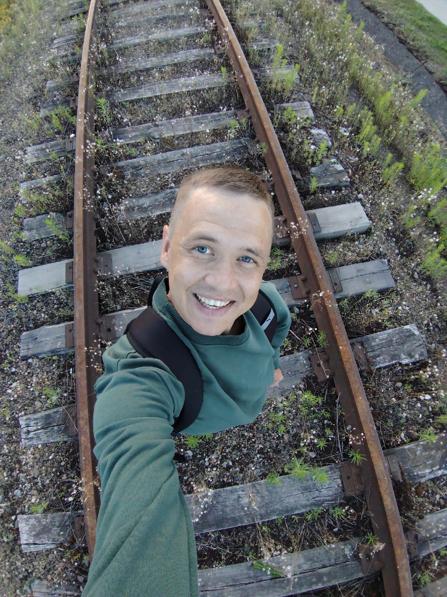 A high-angle, selfie-style shot captures a man standing on a set of abandoned railroad tracks. The man, with short brown hair and bright blue eyes, is smiling widely, his mouth open to reveal white teeth. He is wearing a green long-sleeved shirt and a black backpack. The railroad tracks themselves are weathered, with rusty rails and wooden sleepers overgrown with grass and small white wildflowers. The ballast beneath the tracks consists of gravel and dirt. The scene appears to be outdoors during the daytime, with diffused light suggesting an overcast or early morning/late afternoon setting. The overall mood is cheerful and adventurous, with the man seemingly enjoying his exploration of the disused railway. The location is identified as Chaikovsky, Russia, and while the immediate surroundings show no distinguishing landmarks, the presence of abandoned infrastructure hints at a rural or semi-rural setting. There is no visible text in the image.