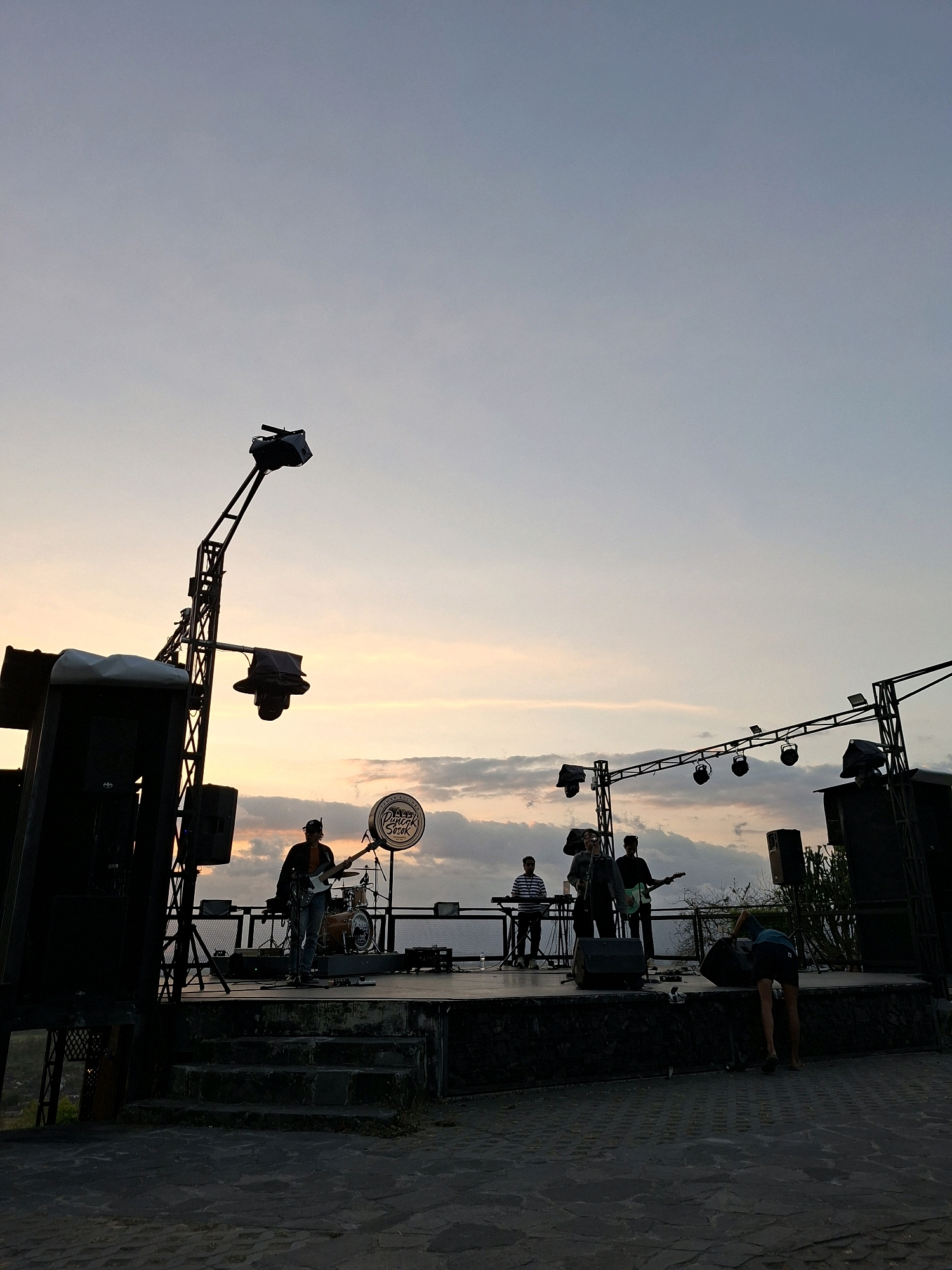 The image depicts an outdoor stage set up for a musical performance at sunset. The sky is filled with soft pastel colors, indicating the twilight hour. On the stage, a band is visible, with several musicians playing instruments and one person standing near a drum set. Large speakers are positioned on either side of the stage, and lighting equipment is suspended above. A circular sign with text is mounted on the stage. In the foreground, a person is bent over, seemingly attending to something on the ground. The setting appears to be an open area with stone paving. The location is identified as Pleret, Indonesia.