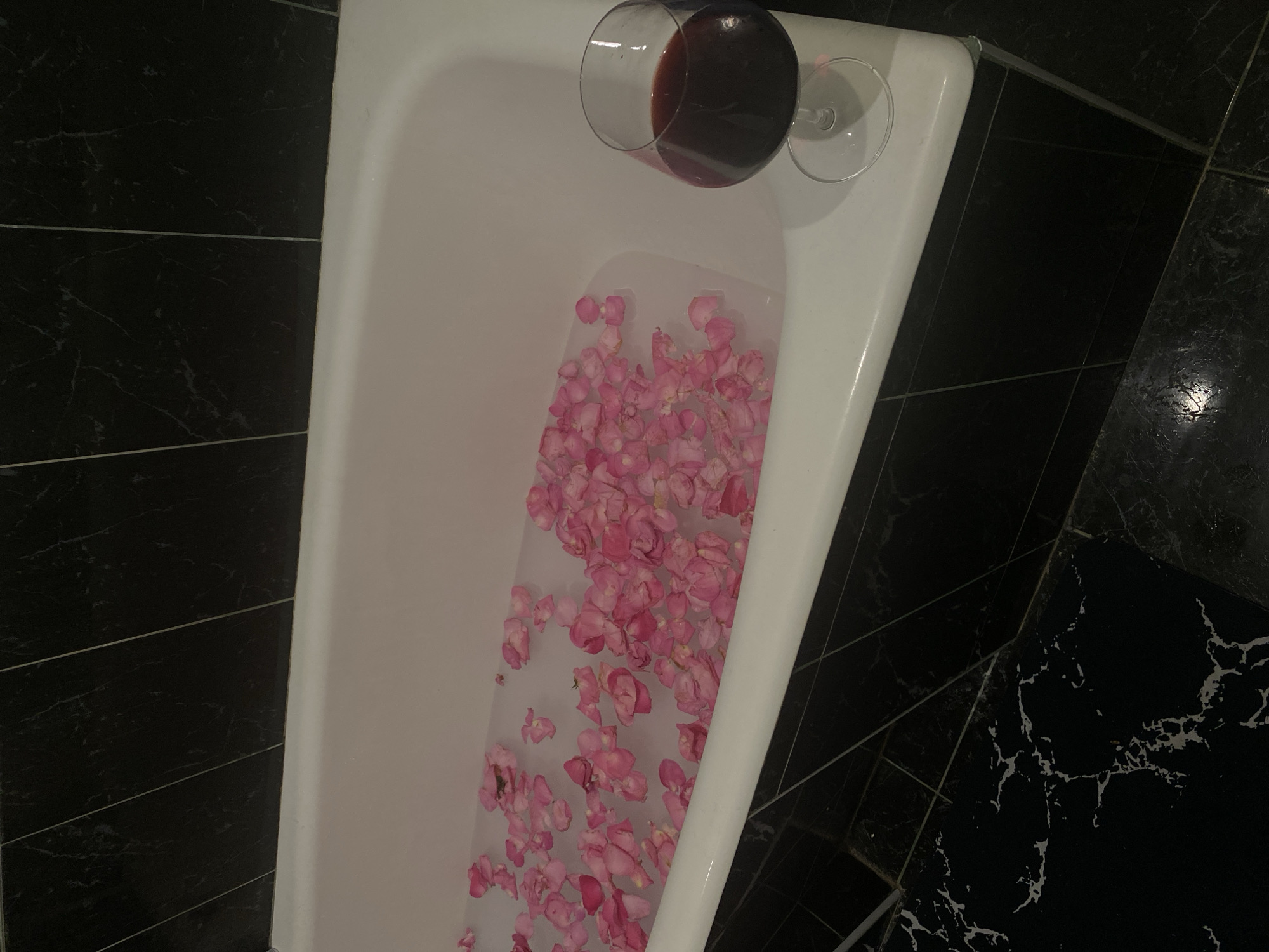 The image shows a white bathtub filled with water and pink rose petals. A half-full glass of red wine is precariously balanced on the edge of the tub. The surrounding tiled walls and floor are black with white veining. There are no people visible in the image. The location is unknown, but not confirmed to be Sochi, Russia.