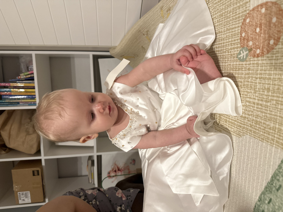 An infant with light blond hair and fair skin is seated upright on a light-colored, patterned mat indoors. The infant wears a white, satin-like dress with short sleeves and a full skirt. The neckline of the dress is embellished with clear and gold-toned beads or pearls. A light-colored tag is visible on the infant's right shoulder. The infant is barefoot and is looking downwards, with both hands touching the fabric of the dress near its feet. The upper left quadrant of the image shows the partially visible left arm and torso of an adult, wearing a grey top with a floral pattern, in close proximity to the infant. The background features a white modular shelving unit with multiple compartments. Visible items on the shelves include a cardboard box, a brown paper bag, a collection of books with colorful spines, and a framed picture of a ballerina. The mat on which the infant sits has a subtle grid pattern and a large, brown-orange strawberry motif in the lower right corner. The scene depicts an indoor residential environment, consistent with a home in the city of Khimki, Russia.