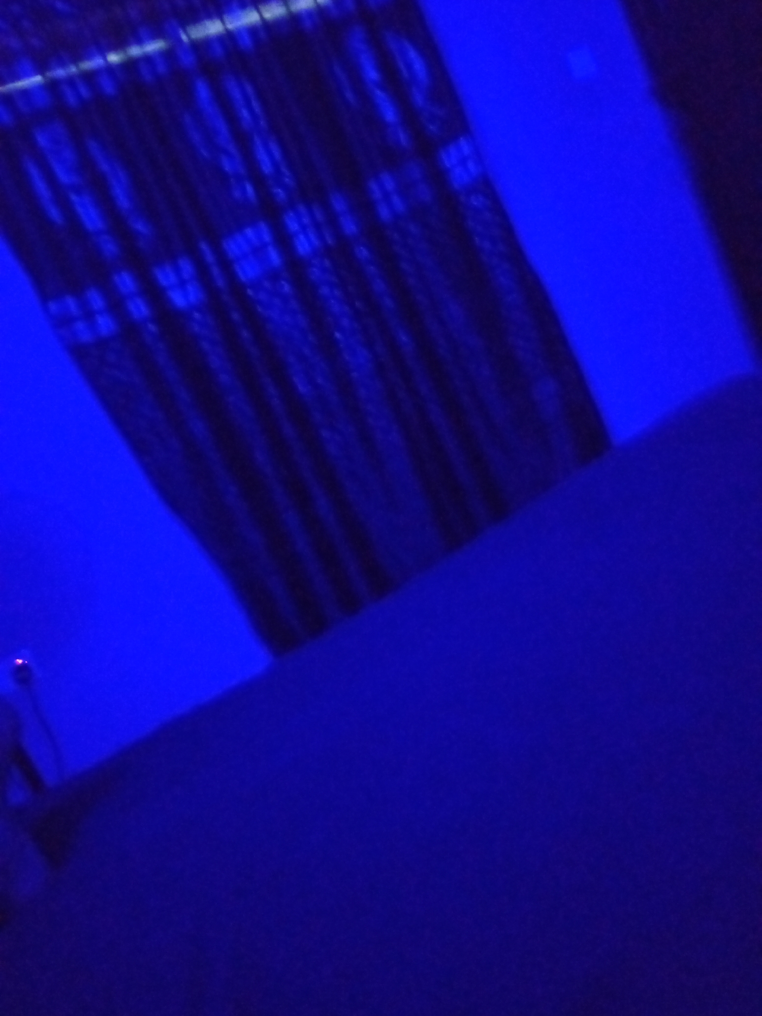 The image displays a dimly lit room bathed in a vibrant blue light. In the upper portion of the frame, a dark, vertically striped curtain is visible, partially obscuring a window or wall. The texture of the curtain suggests it is made of a patterned fabric. A bright blue, untextured surface occupies the lower right and extends upwards, forming a wall or another large surface. The lighting is primarily artificial and casts a strong blue hue over the entire scene. There are no people or discernible actions taking place. The overall environment appears to be indoors, possibly a bedroom or a room designed for ambient lighting. Without further information or visual cues, it is not possible to determine the specific location in Iseyin, Nigeria.