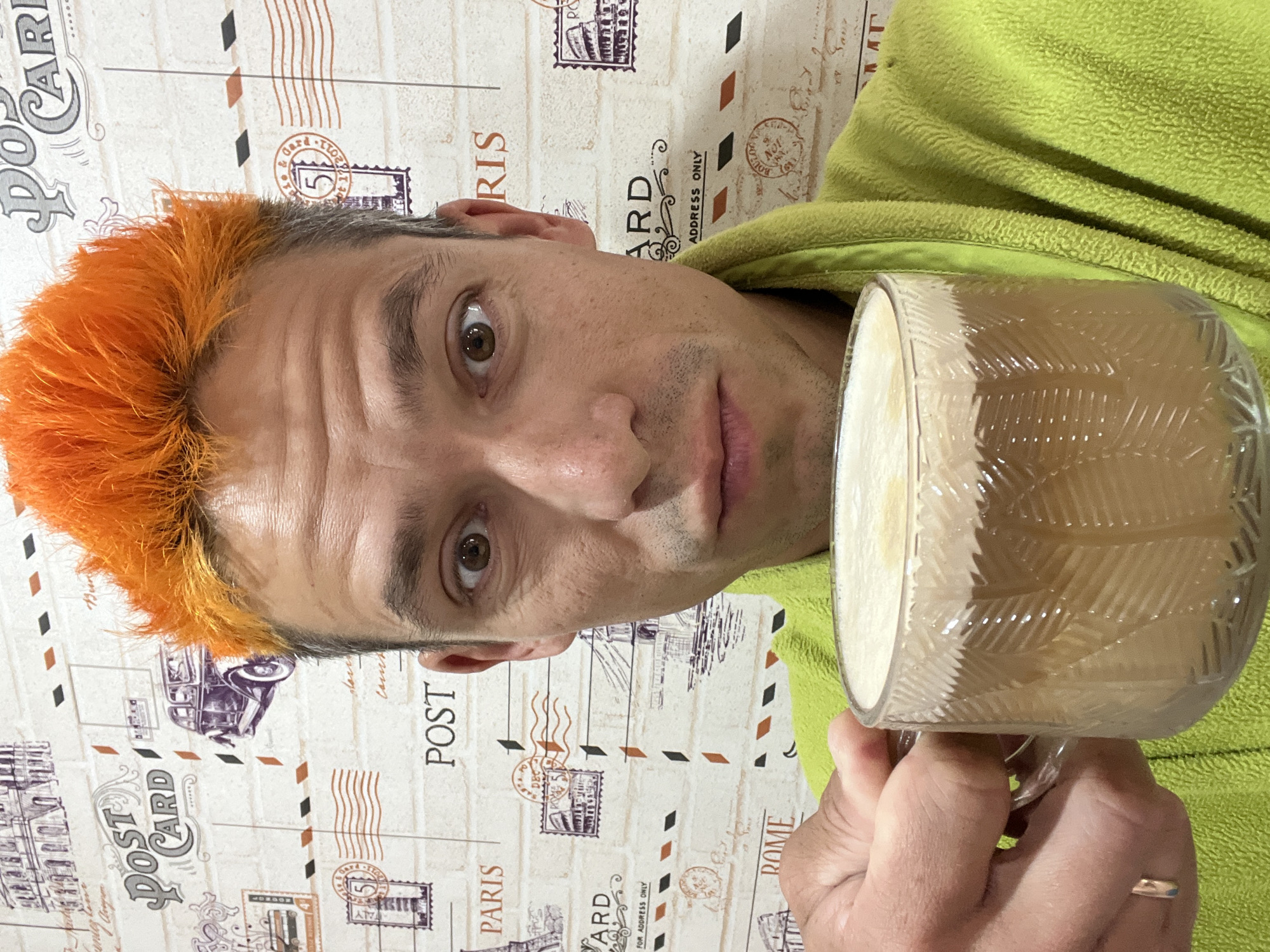 A selfie-style close-up photograph depicts an adult male in an indoor setting, identified as the city of Chaikovsky, Russia. The man occupies the central and lower portions of the frame, looking directly into the camera with wide, brown eyes. He has strikingly bright, spiky orange hair on top, with darker, possibly natural brown or graying roots visible at the temples and sides. His complexion is light, with subtle stubble on his chin and upper lip, and several horizontal lines visible across his forehead. His expression is neutral, with slightly pursed lips.
He is wearing a lime green, textured robe, likely made of fleece or terry cloth, visible on his shoulders and chest. His left hand, with a simple gold-colored band on the ring finger, extends into the lower left of the frame, holding a clear glass mug. The mug features a distinctive molded pattern of vertical, leaf-like or chevron shapes on its exterior. It is filled with a light brown beverage, likely coffee or a latte, topped with a thick layer of creamy-white foam.
The background consists of wallpaper designed to resemble an off-white brick wall covered with vintage travel-themed elements. These elements include various illustrations such as the Eiffel Tower, the Colosseum, a vintage car, and a bridge (possibly Tower Bridge or similar). Text elements like "POST CARD," "POST," "PARIS," "ROME," and "FOR ADDRESS ONLY" are visible. Numerous stylized postage stamps and postmarks are interspersed, some legible with numbers like "5" or "51," and dates such as "17.11.2011," "DEC 2011," and "BOULOGNE NOT 19.11.2010." Wavy and dashed lines, simulating postal markings or airmail stripes, further decorate the background. The lighting is even, suggesting an artificially lit interior.