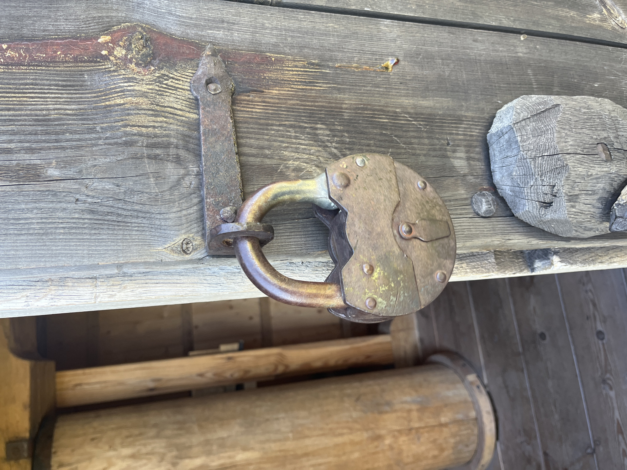 A close-up, eye-level shot focuses on a weathered, oval-shaped padlock made of rusted metal, secured to a rustic wooden door. The padlock's shackle is visible, looped through a rusty metal hasp attached to the door. The door itself is made of aged, vertical wooden planks, with visible grain and signs of wear. To the left of the door, blurred in the background, is a section of a large wooden structure, possibly a barrel or column, and wooden flooring made of horizontal planks. The scene appears to be outdoors, with natural light casting shadows and highlighting the textures of the wood and metal. There are no people present in the image. The overall impression is one of age and historical significance, possibly a detail from a historic building or a traditional structure in Kuressaare, Estonia.
