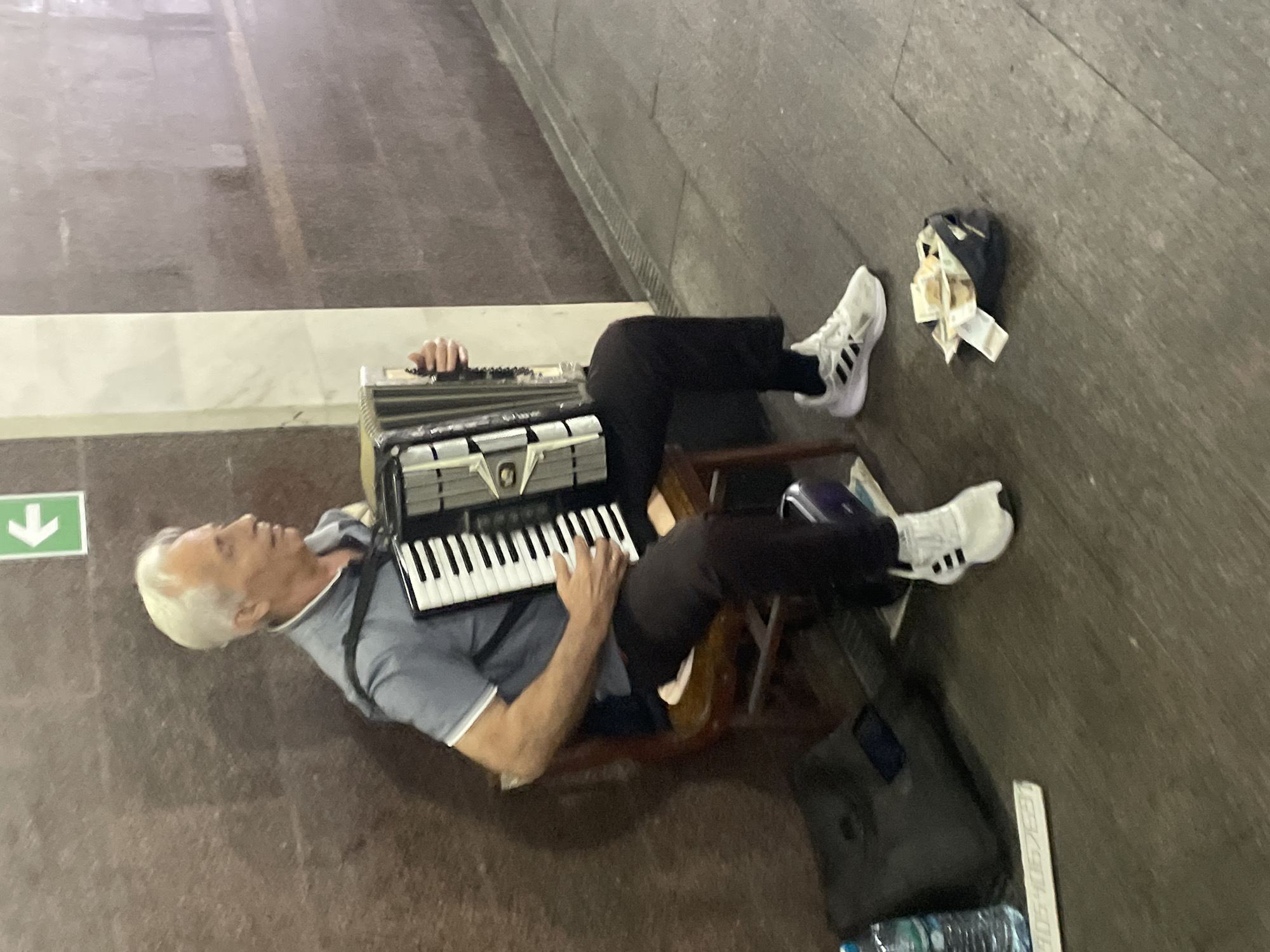 In Sochi, Russia, an elderly man with gray hair, wearing a blue collared shirt and dark pants, sits on a wooden chair playing an accordion. He is positioned in what appears to be an underpass or subway station, indicated by the textured, dark brown walls and the presence of a green sign with a white arrow pointing left. His accordion is black and white, with silver detailing. To his right, a small dark bag lies on the ground filled with cash, suggesting he is busking. In front of the bag, a smartphone is visible. To the left of the musician, a dark bag and a bottle of water are on the ground. The floor is a dull gray concrete, and a metal grate runs along the wall in the background. The scene captures a moment of street performance in an urban setting.