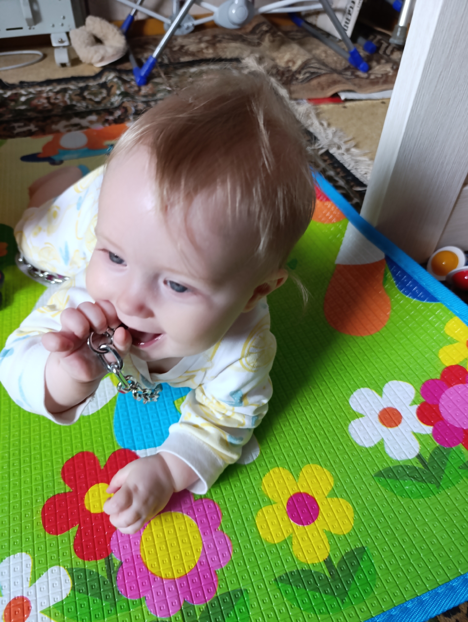 A baby is lying on a colorful play mat with floral patterns. The baby is fair-skinned with blond hair and blue eyes. The baby is wearing a long-sleeved white shirt with yellow and blue designs. The baby is holding and chewing on a metallic toy or object that appears to be a chain with attached small bells. The play mat is green with various colored flower patterns including pink, yellow, and white. In the background, there is a patterned carpet, a set of metal stands with blue accents, and the edge of a white surface. The location is likely Большой Букор, Russia.