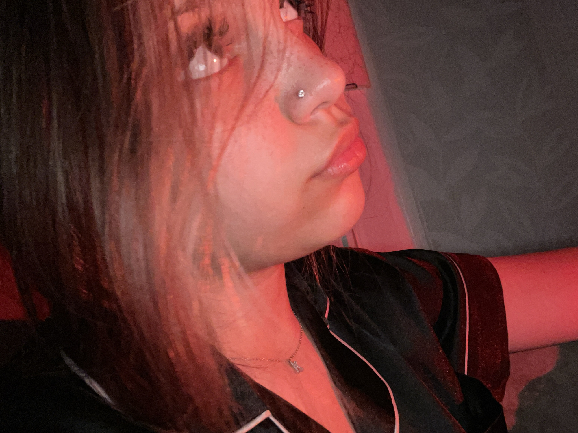 The image shows a close-up of a young person's face, primarily the profile, illuminated by red light. The person has fair skin with some freckles visible on their nose. They have a small, sparkling stud piercing on their right nostril. Their lips are glossy and slightly parted. Eyelashes are prominent, and the eyes are visible, reflecting the red light. The person's hair, which appears to be a light brown or blonde shade, is falling across their face and shoulder. They are wearing a black garment with white trim, possibly pajamas or loungewear, and a delicate silver necklace with a small charm, likely a rabbit, is visible on their neck. The background is dimly lit and appears to be a neutral color, possibly a wall. No specific location in Vaganovo, Russia, can be determined from this image.