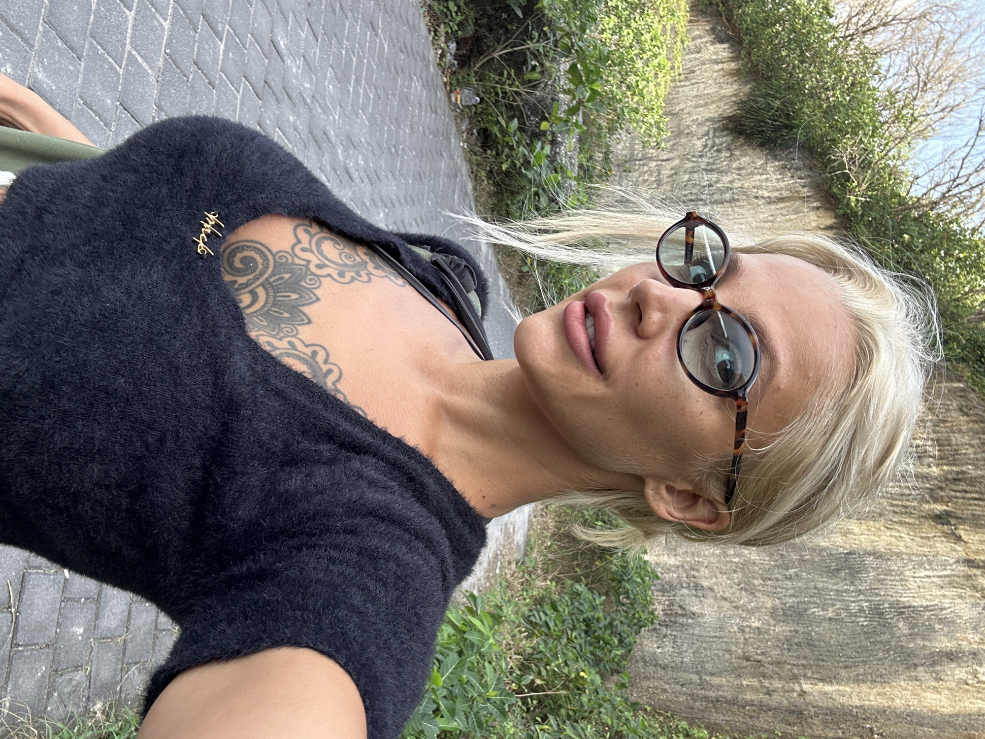 This is a vertical, close-up, outdoor selfie of a person with blonde hair. The subject is wearing a fuzzy black top and round, tortoiseshell-framed glasses. A tattoo is visible on the subject's chest, and a small, gold-colored pin with what appears to be script is attached to the top. The subject's mouth is slightly open, and their expression is neutral.
The background shows a paved walkway made of gray interlocking pavers, with some green foliage and a textured wall to the right. The lighting suggests it is daytime, likely with bright sunlight given the shadows. The location is Kuta Selatan, Indonesia. There is no discernible text in the image.