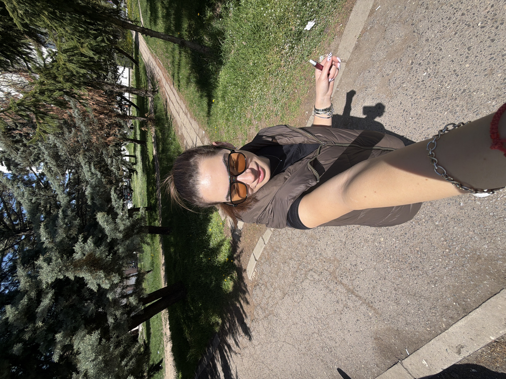 The image is a selfie taken outdoors, featuring a woman from the chest up, looking down at the camera held at arm's length. She is wearing orange-tinted sunglasses, a dark brown sleeveless puffer vest over a black top, and her hair is pulled back. On her left wrist, she wears several bracelets, and she holds a dark red vaping device with her left hand. Her fingernails are painted white with black spots. A silver chain bracelet is visible on her right arm.
The background depicts a sunny outdoor environment, resembling a park or a treelined path. Green grass covers the ground, and various trees, some with dark green needles and others with browner foliage, are visible. A paved path or road runs through the scene, bordered by a concrete curb. Long shadows are cast on the ground by the trees and the person, indicating bright sunshine, likely during mid-morning or late afternoon. A small piece of white debris is present on the paved surface near the curb. The woman exhibits a subtle smile, conveying a relaxed demeanor. No text is visible in the image.