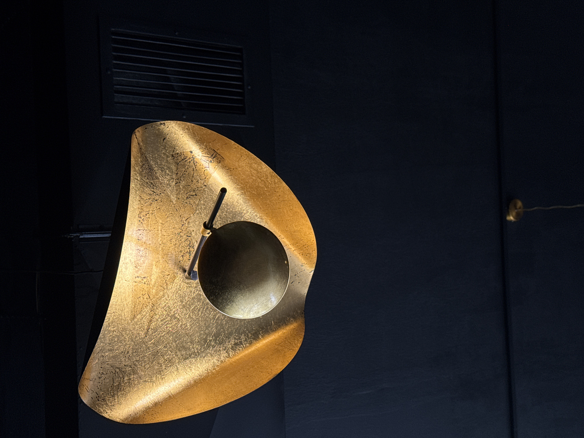 The image features a striking, large, golden, abstract-shaped object, likely a decorative light fixture or a sculpture, set against a dark, minimalist background. The object possesses an organic, irregular form with a visibly textured, metallic surface that appears to be either gilded or made of hammered brass, reflecting light with a warm, uneven glow.
Embedded within the larger golden form, slightly off-center, is a smaller, circular, dark metallic component, which could be a light source or a design element. A slender, dark rod connects this circular part to the main golden structure.
The background is a deep, dark blue or black wall, providing a stark contrast that makes the golden object stand out. In the upper left corner of the background, a dark rectangular vent or grille is visible. On the far right side of the image, barely discernible against the dark wall, is a small, gold-colored, possibly circular element with a thin wire extending from it.
The scene is dimly lit, emphasizing the luminous quality of the golden object. There is no activity taking place; the image captures a static display of modern design. No text is visible in the image. The overall impression is one of contemporary elegance and artistic design, possibly within an upscale interior space like an art gallery, a hotel lobby, or a modern home.