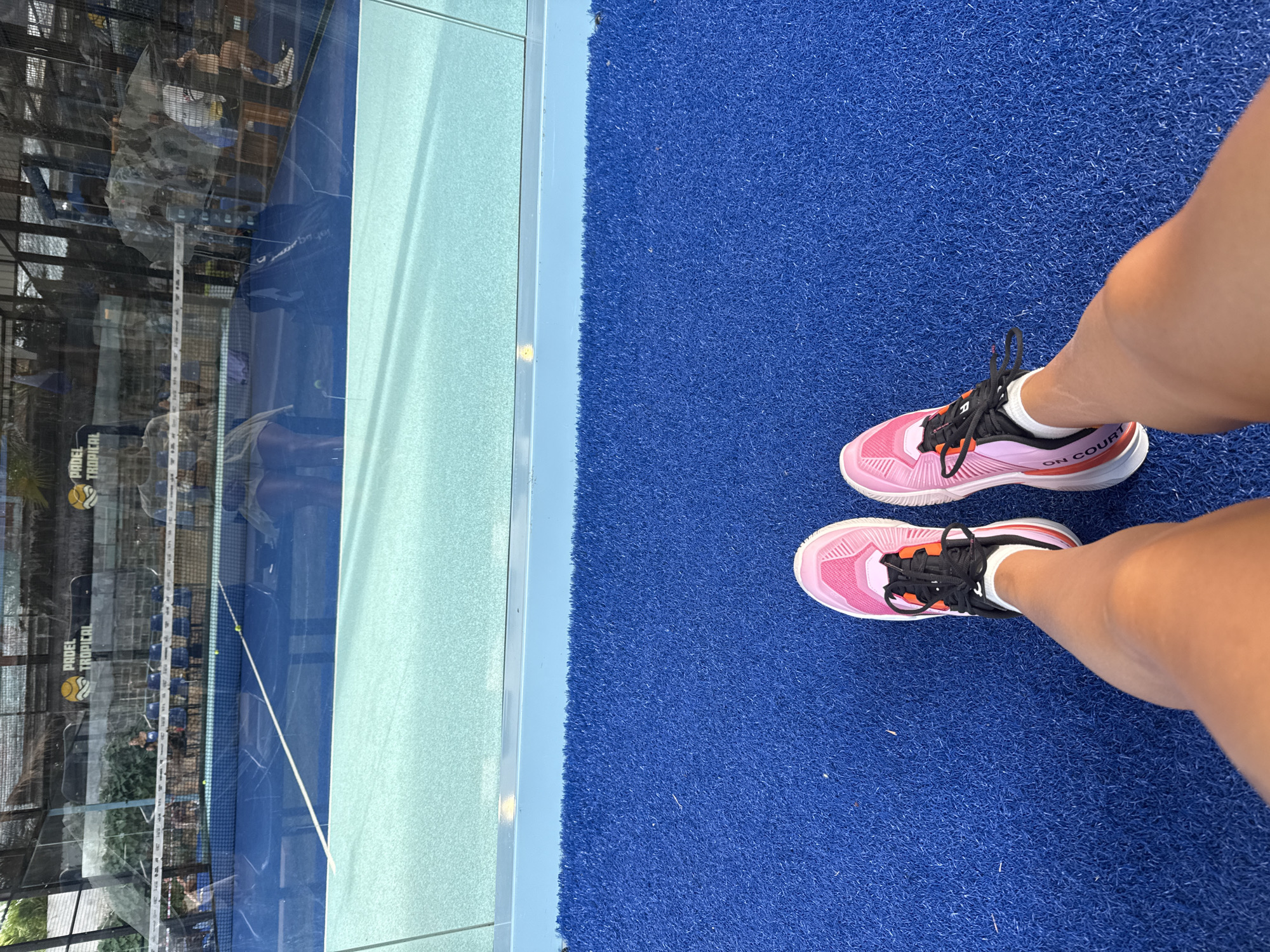 The image shows a first-person perspective of a person's legs and feet, wearing pink and black athletic shoes, standing on a blue carpeted surface. In the background, a reflection in glass reveals a padel tennis court with a blue surface, a net, and people sitting on chairs. Above the court, a sign reads "PADEL TROPICAL". The environment appears to be an outdoor sports facility. The context of Ko Samui District, Thailand is mentioned, suggesting this is likely the location of the Padel Tropical facility.