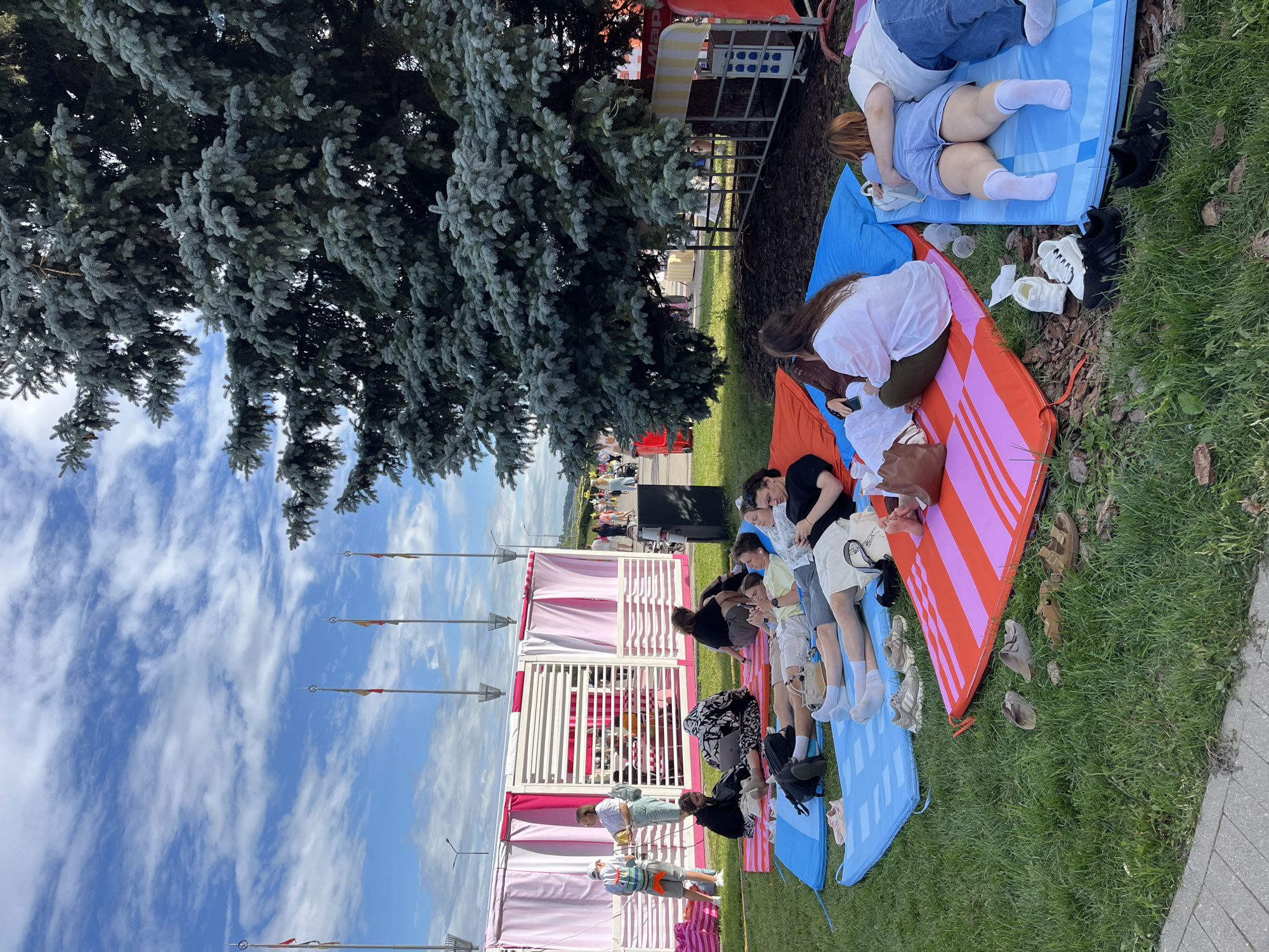 In Nizhny Novgorod, Russia, a group of young adults are relaxing on colorful mats spread across a grassy area, beneath the shade of a large evergreen tree. The people are casually dressed, some lying down and others sitting, engaged in activities like using their phones or conversing. In the background, a white and pink striped structure with open sides suggests a temporary event space or pavilion. Flags on tall poles are visible further in the distance, along with other people walking around. The environment appears to be an outdoor public space, likely a park or festival grounds, on a bright, partly cloudy day.
