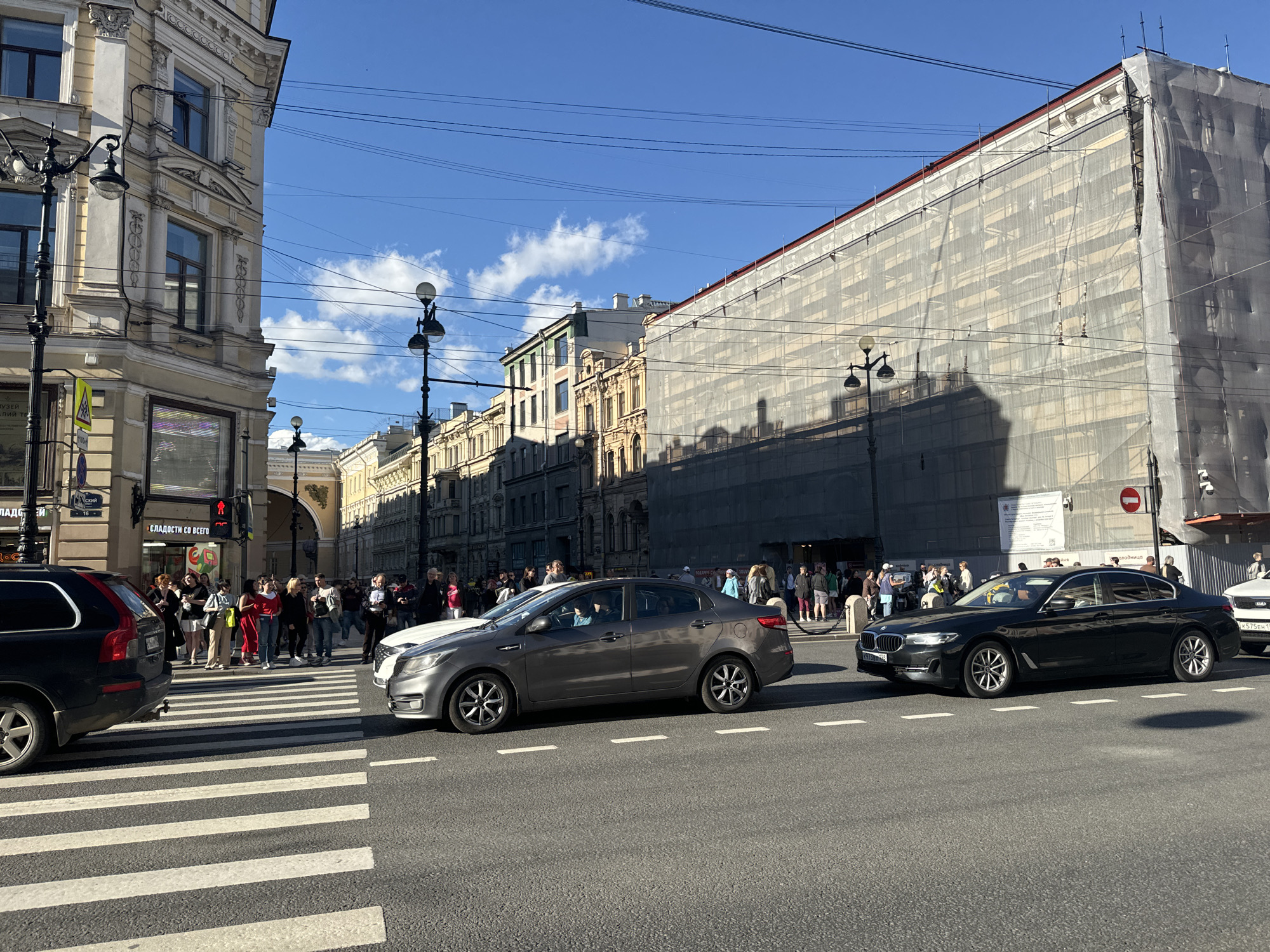This is a daytime, eye-level, medium shot photograph of a street scene in Saint Petersburg, Russia, likely on Nevsky Prospekt due to the architectural style and traffic. In the foreground, a pedestrian crossing with white stripes is visible on the asphalt road. Two cars are in motion over the crossing: a silver sedan in the left lane and a black sedan in the right lane. A black SUV is stopped to the left of the crossing.
On the sidewalk to the left of the frame, a group of pedestrians is gathered, some waiting to cross. They are dressed in casual clothing. The buildings lining the street are multi-story, with ornate architectural details. One building on the right side of the street is covered in white scaffolding and netting. There are streetlights and power lines overhead. The sky is blue with some scattered white clouds. The overall scene depicts a busy urban environment on a sunny day.