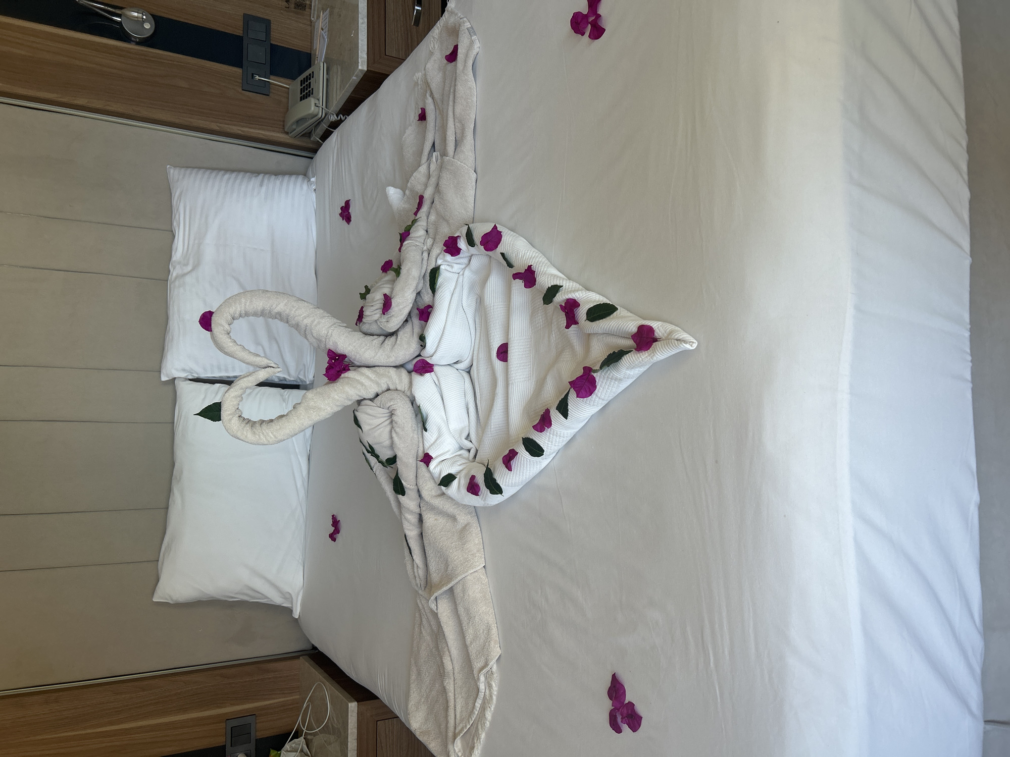 The image depicts a neatly made bed in what appears to be a hotel room, likely located in Didim, Turkey. The bed is covered with white linens and features two towel swans arranged in a heart shape. Pink flower petals and green leaves are scattered artistically around the towel sculptures and on the bed. Two white pillows are positioned at the head of the bed. To the left and right of the bed, bedside tables are visible, each with a lamp and a phone. The overall impression is one of romantic or welcoming accommodation.