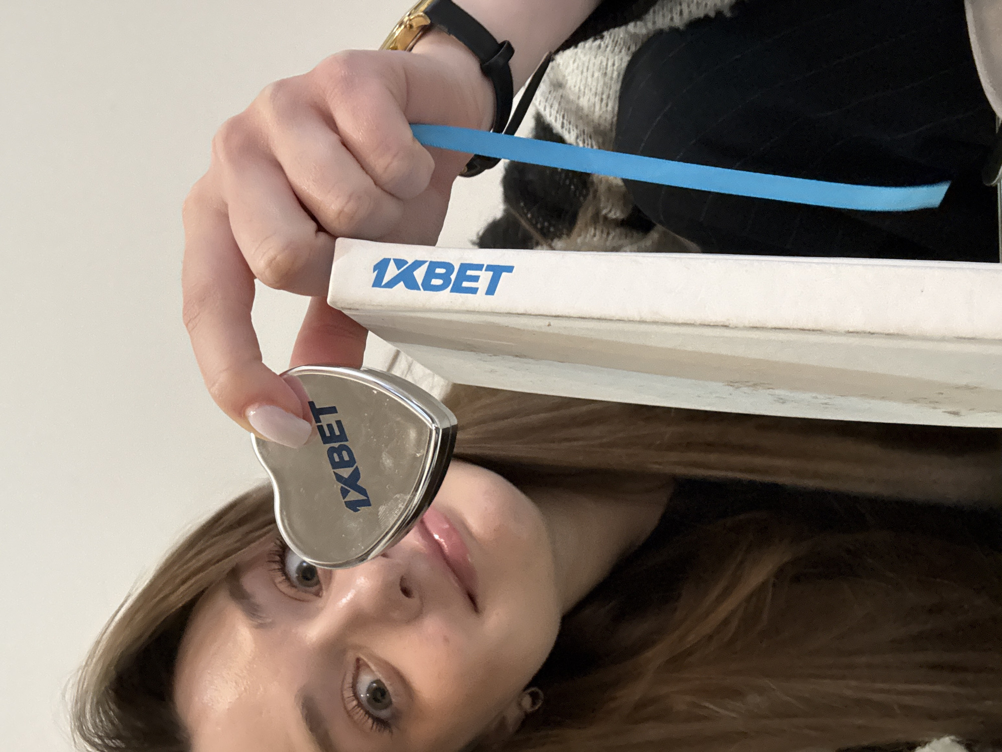 A woman with long brown hair is holding a heart-shaped object and a white rectangular object with the word "1XBET" printed on it. The woman's face is visible below the heart-shaped object, showing her eyes, nose, and mouth. Her left hand holds the heart-shaped object, while her right hand holds the white rectangular object, with a blue ribbon placed on top of it. A watch is visible on her right wrist. The woman's clothing is partially visible, showing a patterned sweater and a black pinstripe garment. The location in the city of Kragujevac, Serbia, cannot be determined from the image.