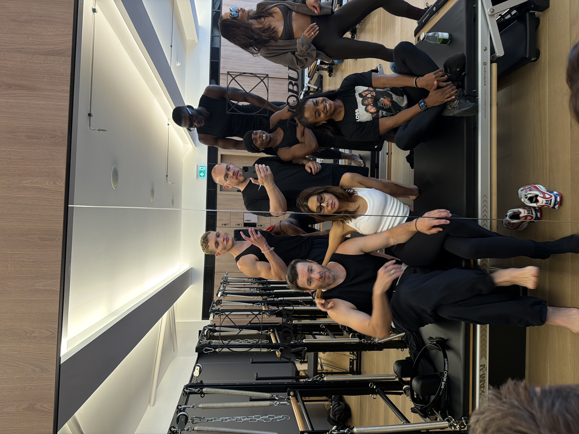The image, taken in a Pilates studio in London, United Kingdom, shows a group of eight individuals posing for a mirror selfie. The studio features a polished wooden floor, wood-paneled walls, and rows of Pilates reformers. The group consists of four men and four women, all dressed in athletic wear. Most of the individuals are smiling or looking at the camera. One man, in the foreground, is seated on a Pilates reformer, his arm around a woman sitting next to him. Another man, standing behind them, is holding a phone and taking the photo. In the background, other members of the group are visible, some standing and some sitting. The lighting in the studio is bright, with overhead track lighting and natural light filtering in from unseen windows. A large mirror reflects the group and the studio environment. The overall atmosphere appears to be casual and friendly, suggesting a post-workout gathering or a group class.