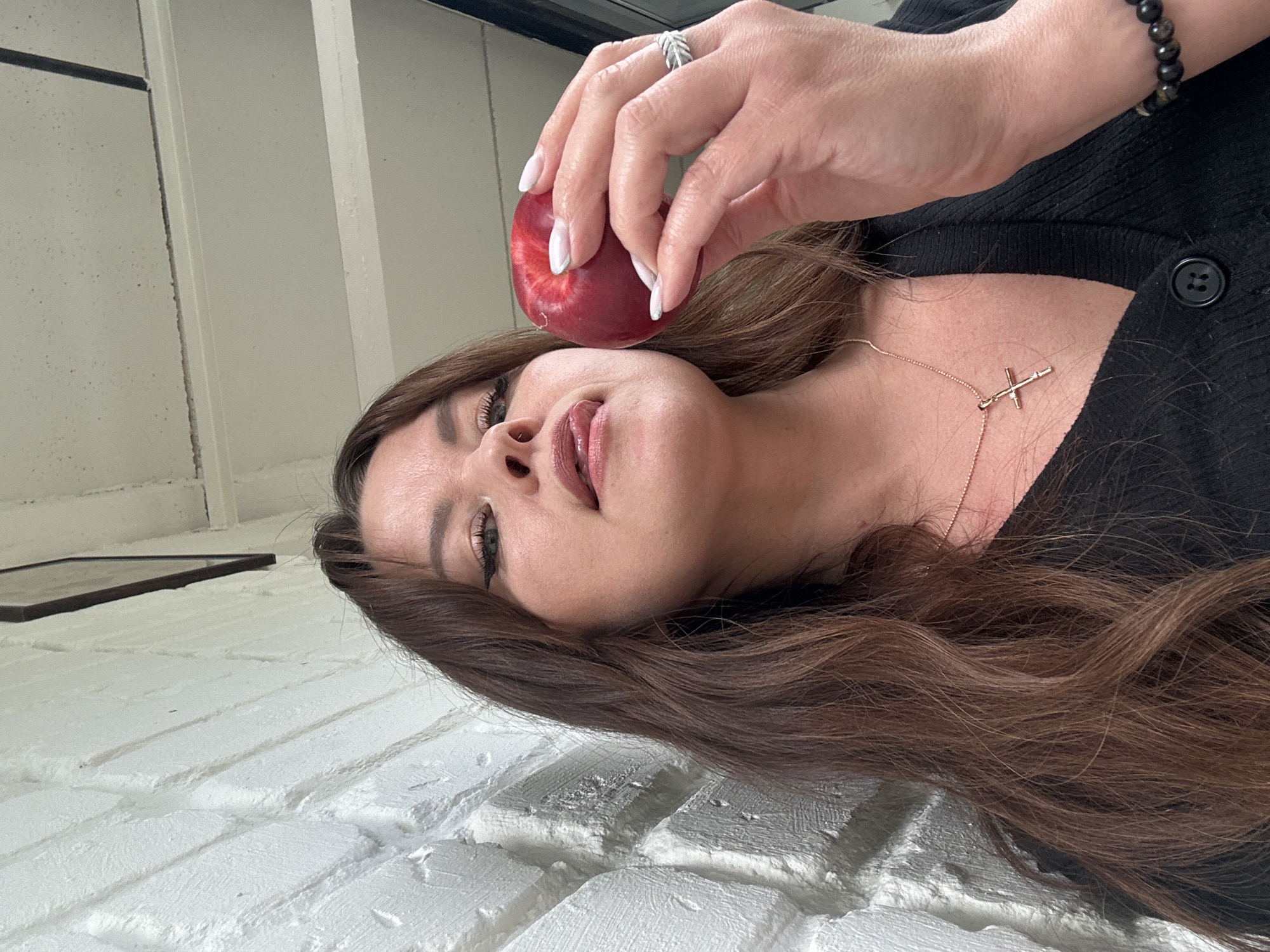 The image shows a woman with long brown hair and makeup, holding a red apple in her right hand. She is wearing a black top and a necklace with a cross pendant. The background is a white brick wall and a white ceiling with dark beams. The image is a close-up of the woman's face and upper body. It is not possible to determine the location in the city of Chaikovsky, Russia from this image.
