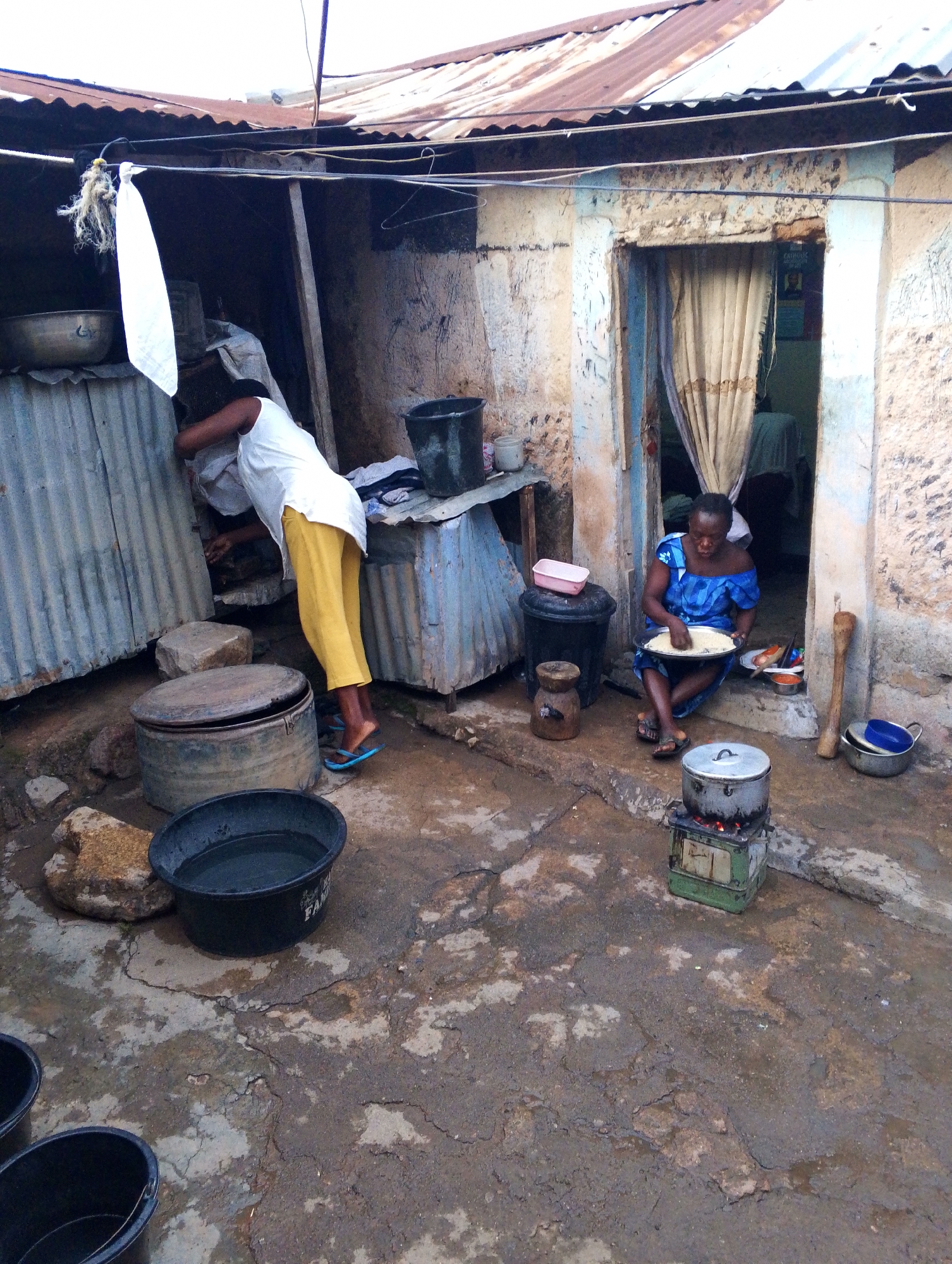 The image depicts an outdoor scene in Jos, Nigeria, likely a courtyard or yard adjacent to a dwelling. Two women are visible. One woman is standing and appears to be interacting with corrugated metal sheeting in a lean-to structure; she is wearing a white top and yellow trousers. The other woman is sitting in the doorway of a mud-brick building, wearing a blue dress and holding a white plate. A pot is cooking on a small stove in front of her. Several buckets, bowls, and containers are scattered around the courtyard. The ground is cracked and appears to be earth or concrete. The structures have corrugated metal roofs. The overall scene suggests a domestic environment, possibly a kitchen and living space.