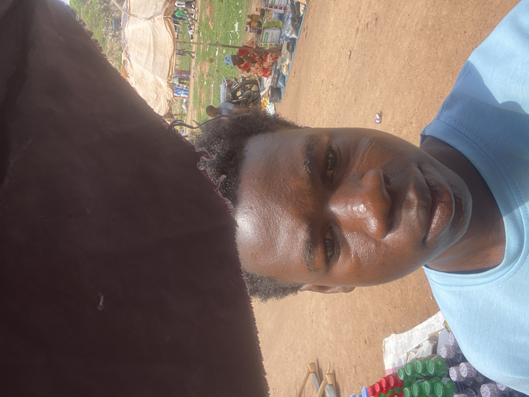 The image, presented as a sideways portrait, features a close-up selfie of a person with dark skin, wearing a light blue t-shirt. Their face, illuminated by bright sunlight, exhibits a natural sheen, and they have short, dark, curly hair. They look directly at the camera with a neutral expression. A large portion of the left foreground is covered by a dark brown fabric or garment.
The background reveals a bustling outdoor market scene, characteristic of Mararaba, Nigeria. The ground is unpaved earth, bathed in clear daylight, indicating a sunny day. Numerous blurred figures are scattered throughout, engaged in various market activities. Several makeshift market stalls, some with light-colored tarp coverings, are visible. One prominent stall in the mid-ground shows a person in a red and white patterned outfit with a blue head covering, seated behind a display of goods including bottles and packaged products. A motorcycle is parked near a stall. In the immediate foreground, next to the main subject, are stacks of green-capped and white-capped bottles, alongside wooden objects resembling tools. Faint, unreadable text, possibly "NATE," can be seen on some distant product labels. The scene conveys the busy atmosphere of a local marketplace.