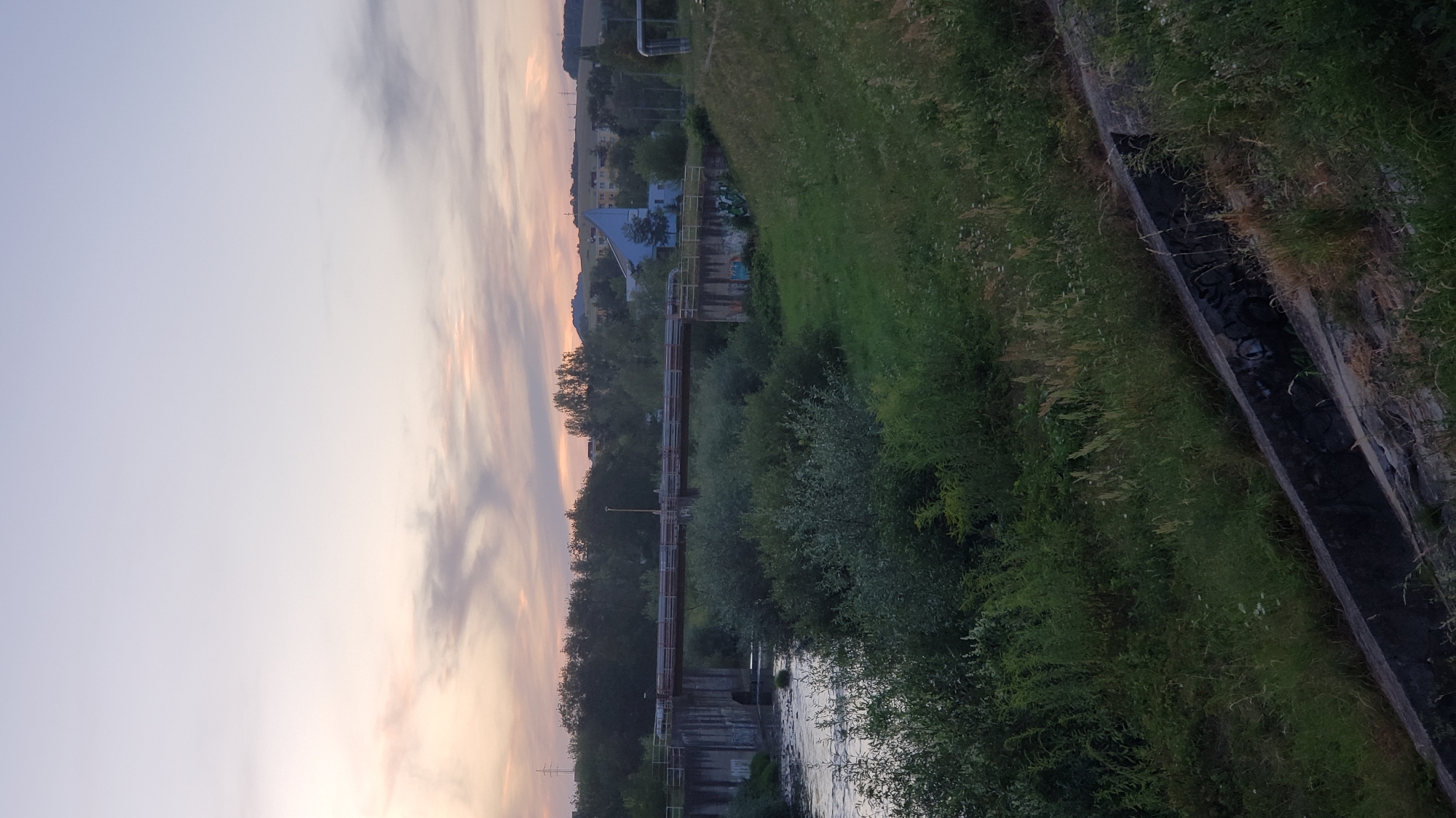 The image shows a wide shot of a river, a bridge, and surrounding greenery in Prešov, Slovakia. The sky is a pale lavender with wisps of pink and orange clouds, suggesting it is either sunrise or sunset. The bridge, made of concrete and metal, spans the river, which appears to be flowing calmly. Dense green foliage, including trees and bushes, covers the riverbanks and extends into the background. A modern building with a distinct blue, angular roof is visible on the right side of the frame, behind the bridge and surrounded by more trees. The foreground features a grassy embankment sloping down towards a concrete structure, likely part of a drainage system, with some graffiti visible on its side. The overall scene depicts a somewhat industrial yet natural setting within the city.