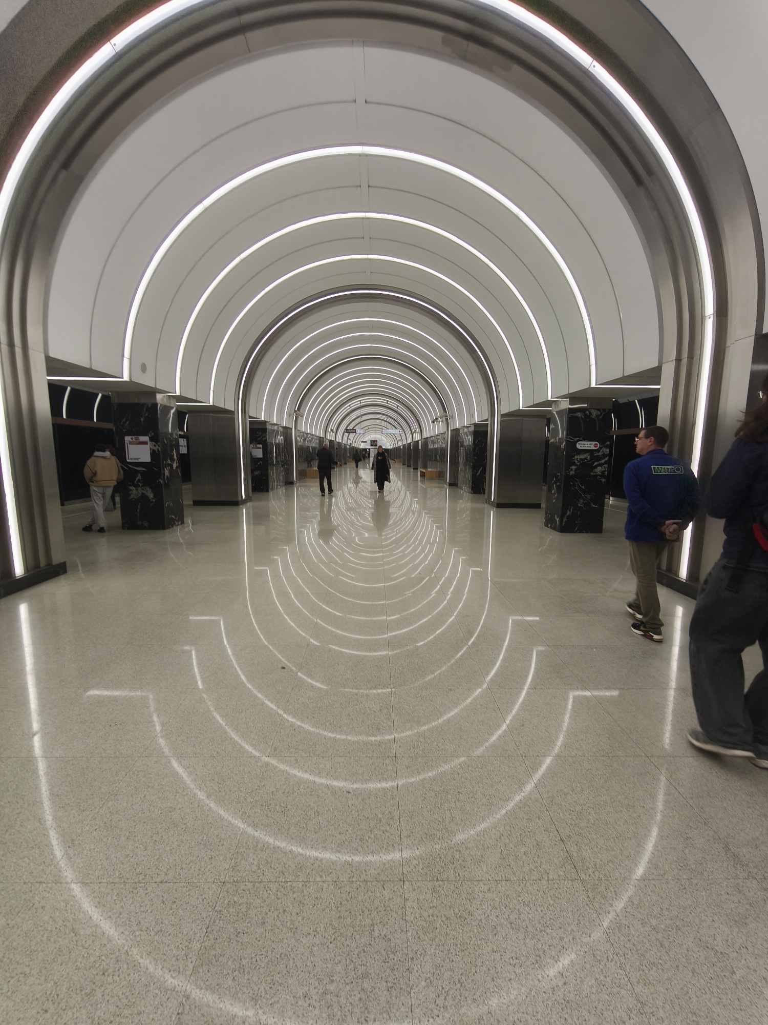 The image depicts the interior of the CSKA metro station in Moscow, Russia. The station has a distinctive architectural design with a series of arched, white-lit tunnels. The floor is a light-colored terrazzo. People are walking through the station. There are pillars or structures clad in dark marble or a similar material along the sides of the walkway. The overall atmosphere is modern and clean.