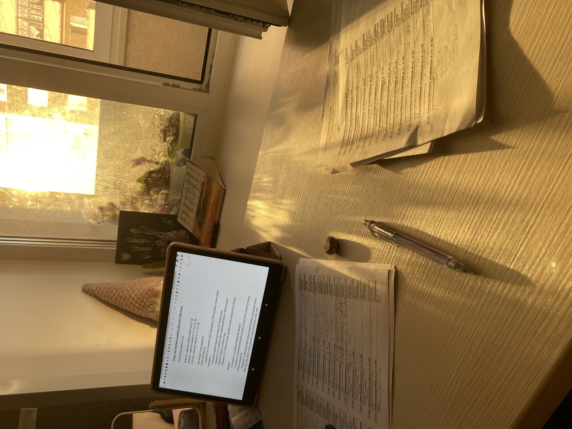 A top-down shot reveals a desk bathed in warm sunlight streaming through a window. The window offers a glimpse of buildings across the way, potentially placing the scene in an urban setting. On the desk, a tablet displays text in Russian, suggesting it's being used for study or work. To the right of the tablet, papers filled with mathematical equations and notes are scattered. A pen lies next to these papers, indicating active engagement with the material. A small, round object, perhaps a cork or eraser, sits nearby. The desk itself appears to be made of a light-colored wood. Based solely on the content of the documents, the setting is likely in Russia, but no specific information is available to pinpoint the exact location in Votkinsk.