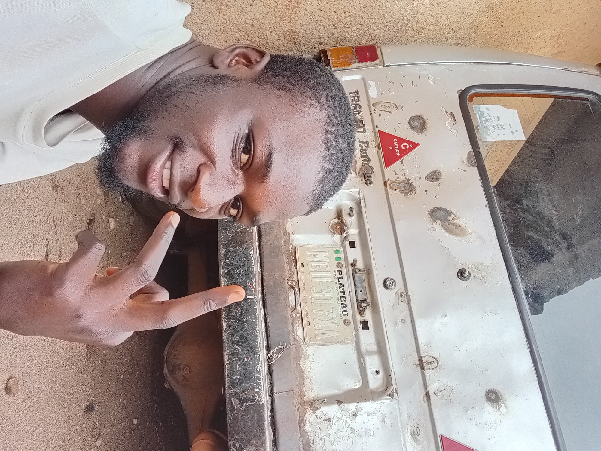 In Bukuru, Nigeria, a young man with dark skin, a beard, and a white t-shirt poses for a selfie. He is positioned in the foreground, his head and upper body taking up the left half of the frame. He is smiling and holding up two fingers in a peace sign with his right hand. Behind him is the rear of a silver van. The van's rear is marked with several dents and patches of rust, indicating wear and tear. A red triangular "CAUTION" sticker is visible on the back of the van. A Nigerian license plate reading "MGU-317XA" is also attached to the rear bumper. The ground beneath the van is a mixture of dirt and gravel.