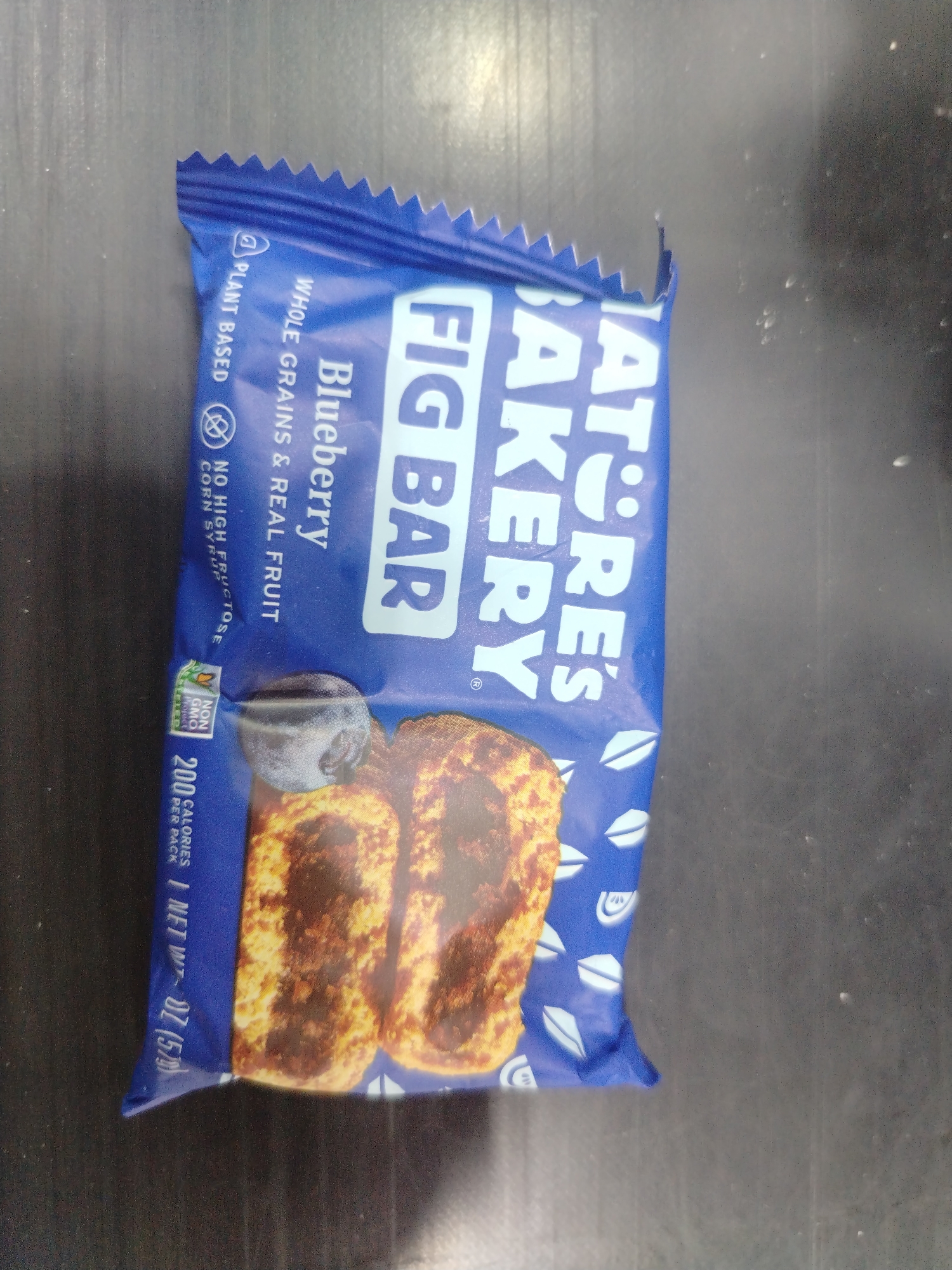 The image shows a close-up, overhead shot of a single Nature's Bakery Fig Bar package. The package is predominantly blue with white lettering and graphics. The text on the package includes "NATURE'S BAKERY," "FIG BAR," "Blueberry," "WHOLE GRAINS & REAL FRUIT," "PLANT BASED," and nutritional information stating "200 CALORIES PER PACK" and "1 NET WT - 1.85 OZ (52g)". There are also icons indicating "NON GMO PROJECT VERIFIED" and a prohibition symbol over "HIGH FRUCTOSE CORN SYRUP". The package is oriented vertically and slightly angled. Two fig bars are visible through a transparent portion of the packaging at the bottom, showing a textured, baked appearance. The background is a dark, textured surface, possibly a countertop or table. There are no people or any discernible elements that place this scene specifically in Juan Griego, Venezuela.