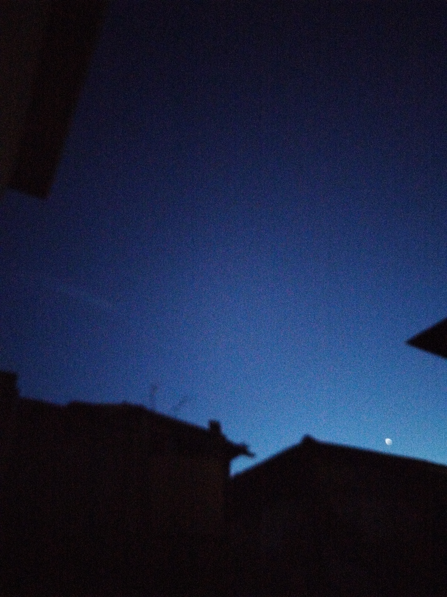 The image shows a dark blue sky with a faint crescent moon visible on the right side. Silhouetted against the sky are the rooftops of several buildings, suggesting an urban or town setting. The buildings appear to be of a traditional architectural style, possibly residential. The overall impression is that of twilight or early night. The location is specified as Smolyan, Bulgaria.