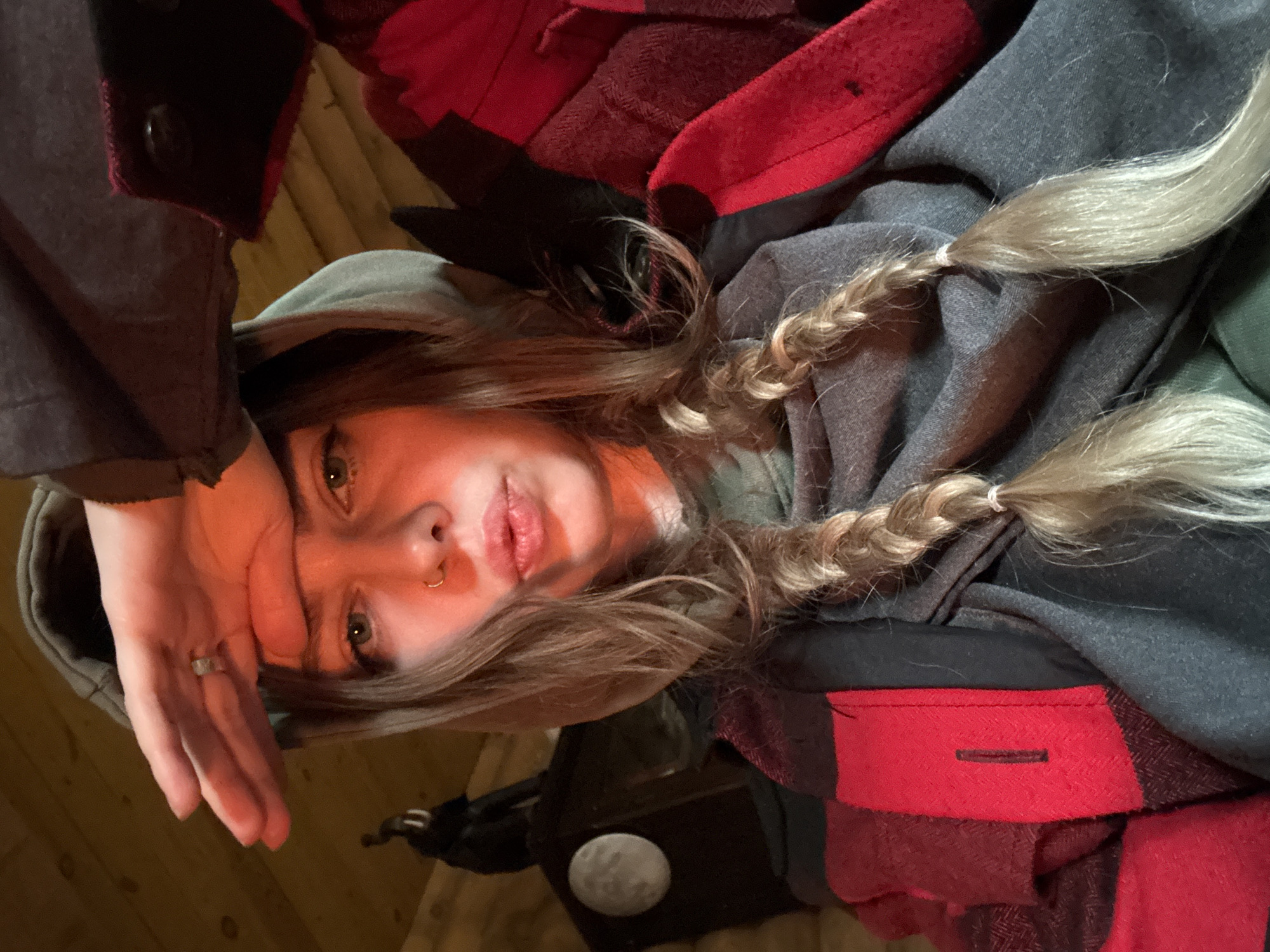 A young person with long, light-colored hair, styled in two braids, is depicted in a close-up, upward-facing shot, appearing to be lying down. They are looking directly at the camera with a direct and relaxed expression, their lips slightly parted. A small hoop piercing is visible in their left nostril, and their eyes are light-colored.
The person is wearing a grey hooded top, partially covered by a red and black plaid jacket or shirt. Their left hand is resting on their forehead, revealing a ring on their left ring finger. The braids are secured with white hair ties.
The background consists of warm-toned wooden floorboards, indicating an indoor setting, likely a room or cabin. In the lower part of the image, a dark object, possibly furniture or a bag, supports a small, spherical white object that resembles a miniature moon or a decorative orb. The lighting is warm and somewhat dim, suggesting an evening or low-light indoor environment. No text is visible in the image. The attire and casual posture contribute to a cozy and personal atmosphere.
