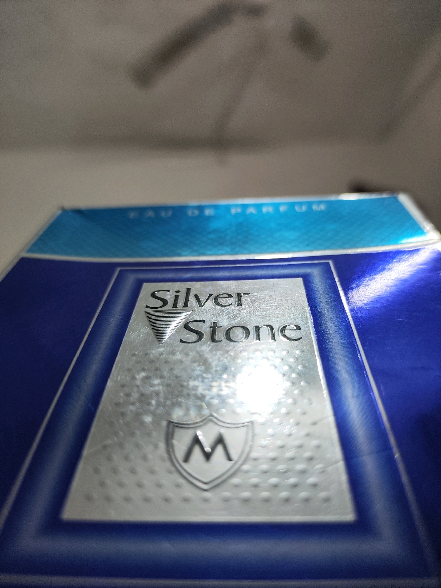 The image is a close-up, angled shot of a product box for "Silver Stone" eau de parfum. The box is primarily blue with a silver rectangular label in the center. The label has the words "Silver Stone" printed in a metallic font, with a triangular emblem above "Stone." Below this, there is a shield-like emblem containing the letter "M." The top edge of the box features a blue strip with white text that reads "EAU DE PARFUM." The background appears to be a ceiling with a fan or light fixture, suggesting the photo was taken indoors. The scene depicts a product for sale, likely in a retail environment or at home. There are no people or specific landmarks visible that would indicate a location in Aligarh, India.