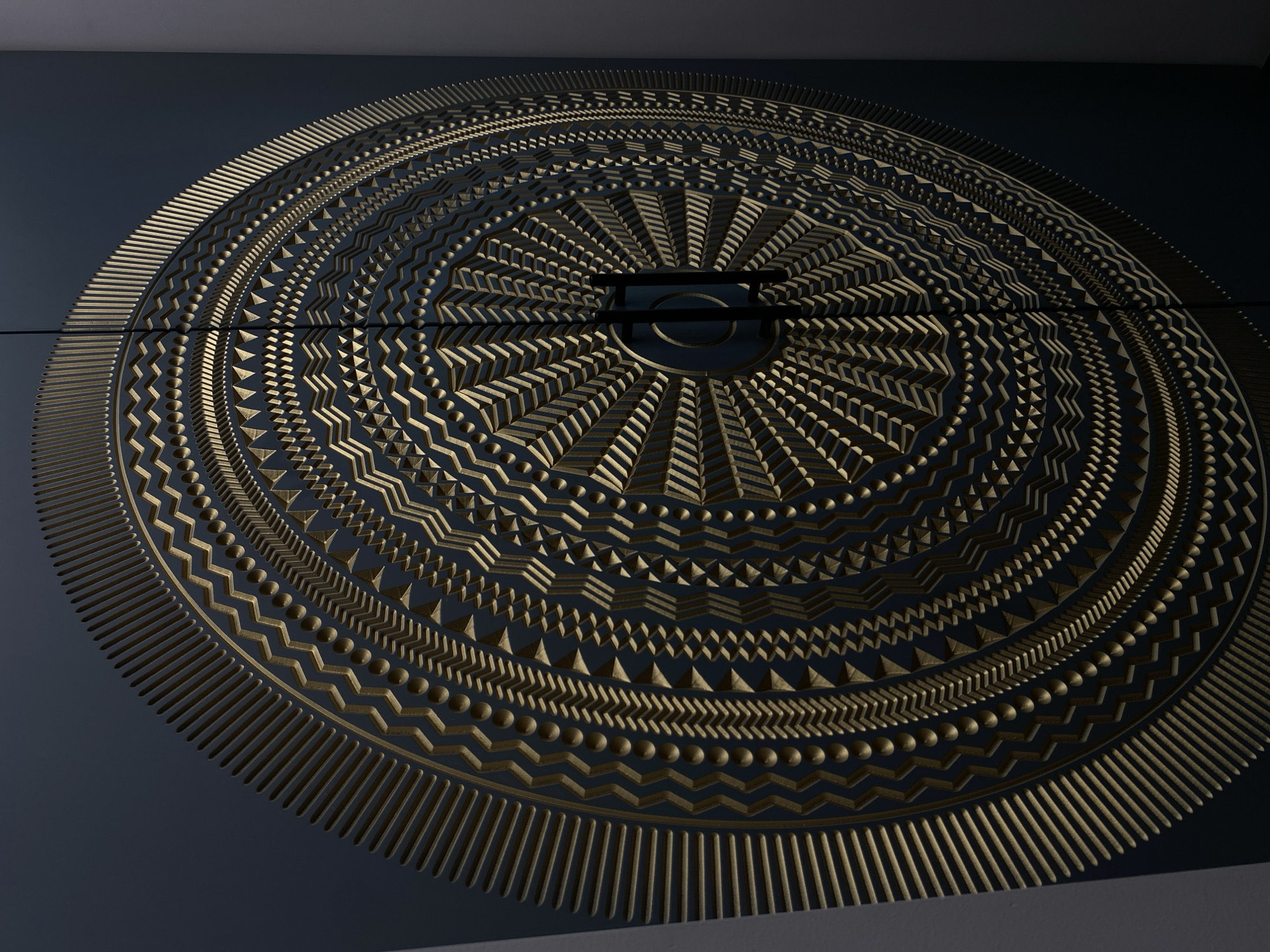The image showcases a large, circular, ornamental design carved into a dark, possibly wooden or composite, surface. The design consists of concentric rings featuring various geometric patterns, including lines, zigzags, triangles, and dots. A handle is visible in the center of the design, suggesting it might be a door or a cabinet feature. There are fine dark lines running across the center of the circle which suggests it is comprised of two or more pieces.
The location in Votkinsk, Russia, is not directly discernible from the visual content. There is no way to determine the location from the image.