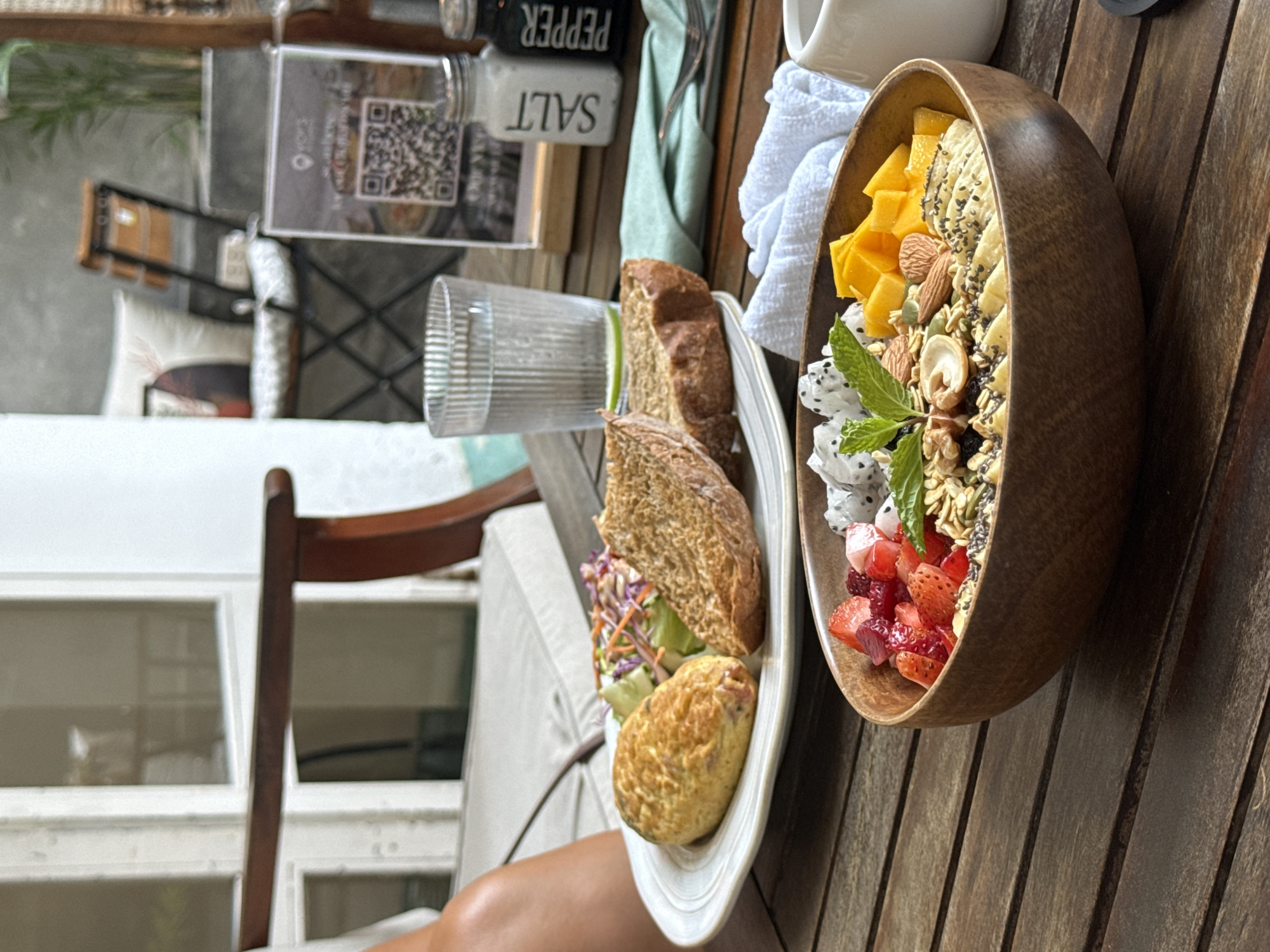A tabletop breakfast scene on a wooden deck in Đà Nẵng, Vietnam. A wooden bowl filled with chopped mangoes, dragon fruit, strawberries, bananas, nuts, and granola is on the right. A white plate containing two slices of bread, a small salad, and a pastry is on the left. A glass of water, salt and pepper shakers, a white cloth, and a white mug are also present. A chair and part of a person's leg are visible in the foreground. A menu with a QR code is present. The background shows a glimpse of an outdoor setting.