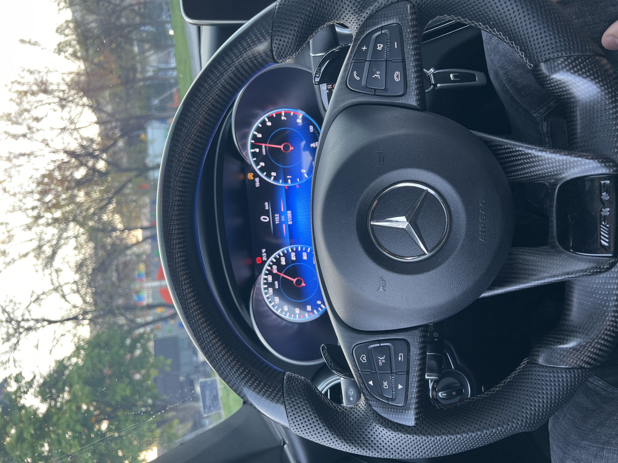 Here's a detailed description of the image:
**Scene:** The image depicts the interior of a Mercedes-Benz vehicle. The focus is on the dashboard and steering wheel.
**Objects:**
* **Steering Wheel:** Carbon fiber and perforated leather steering wheel with Mercedes-Benz logo at the center. Control buttons are visible on both sides of the steering wheel. A car key is visible.
* **Dashboard:** A digital dashboard displays information such as speed (0 km/h), mileage (81088 km).
* **Exterior View:** The windshield provides a view of the outside environment, including trees.
**Environment:**
*The exact location within Plovdiv, Bulgaria cannot be determined from the available visual information.*