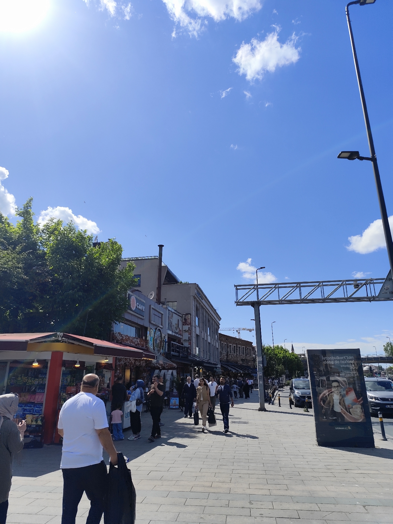 The image captures a bustling street scene in an urban environment. Pedestrians are walking along a paved sidewalk, with a mix of casual clothing and headscarves. A variety of shops and restaurants line the street, including a cafe with outdoor seating and a kiosk with merchandise. An advertisement board is visible on the right side of the image, and cars are parked along the street. The sky is bright blue with scattered clouds, and sunlight illuminates the scene. A metal framework spans the street overhead, and a tall streetlight stands on the right.