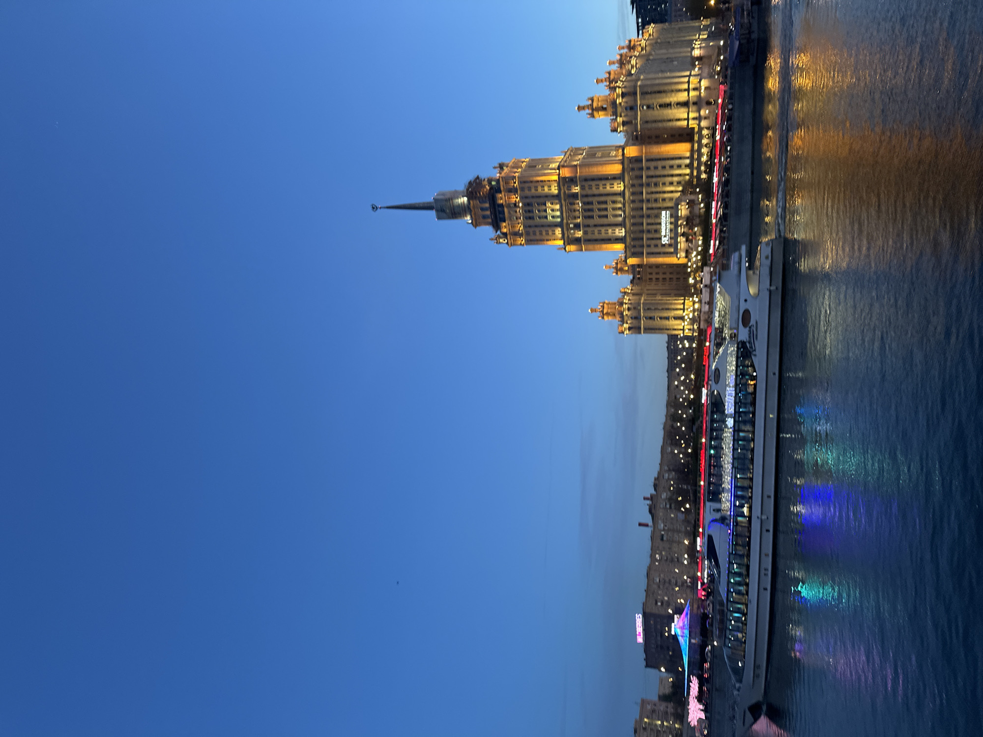 The image presents an urban waterscape captured at dusk or early night. A grand, ornate skyscraper, identifiable as the Radisson Collection Hotel, Moscow (formerly Hotel Ukraina), dominates the right side of the frame. The building is extensively illuminated with warm, golden lights, highlighting its architectural details and prominent spire against a deep, clear blue sky.
Alongside the building runs a wide river, likely the Moskva River, reflecting the building's golden glow and the vibrant, multi-colored lights from a large passenger boat. This boat is brightly lit with red, blue, green, and purple lights, casting colorful reflections onto the calm water. Faintly visible text on the boat includes "Radisson".
Across the river, other city buildings are present in the background, also illuminated, contributing to the urban night scene. The sky transitions from a lighter blue near the horizon to a darker shade overhead, indicative of twilight.
Visible text on the main building reads "RADISSON COLLECTION" and "HOTEL MOSCOW". The overall atmosphere is one of serene urban beauty and grandeur during the evening hours.