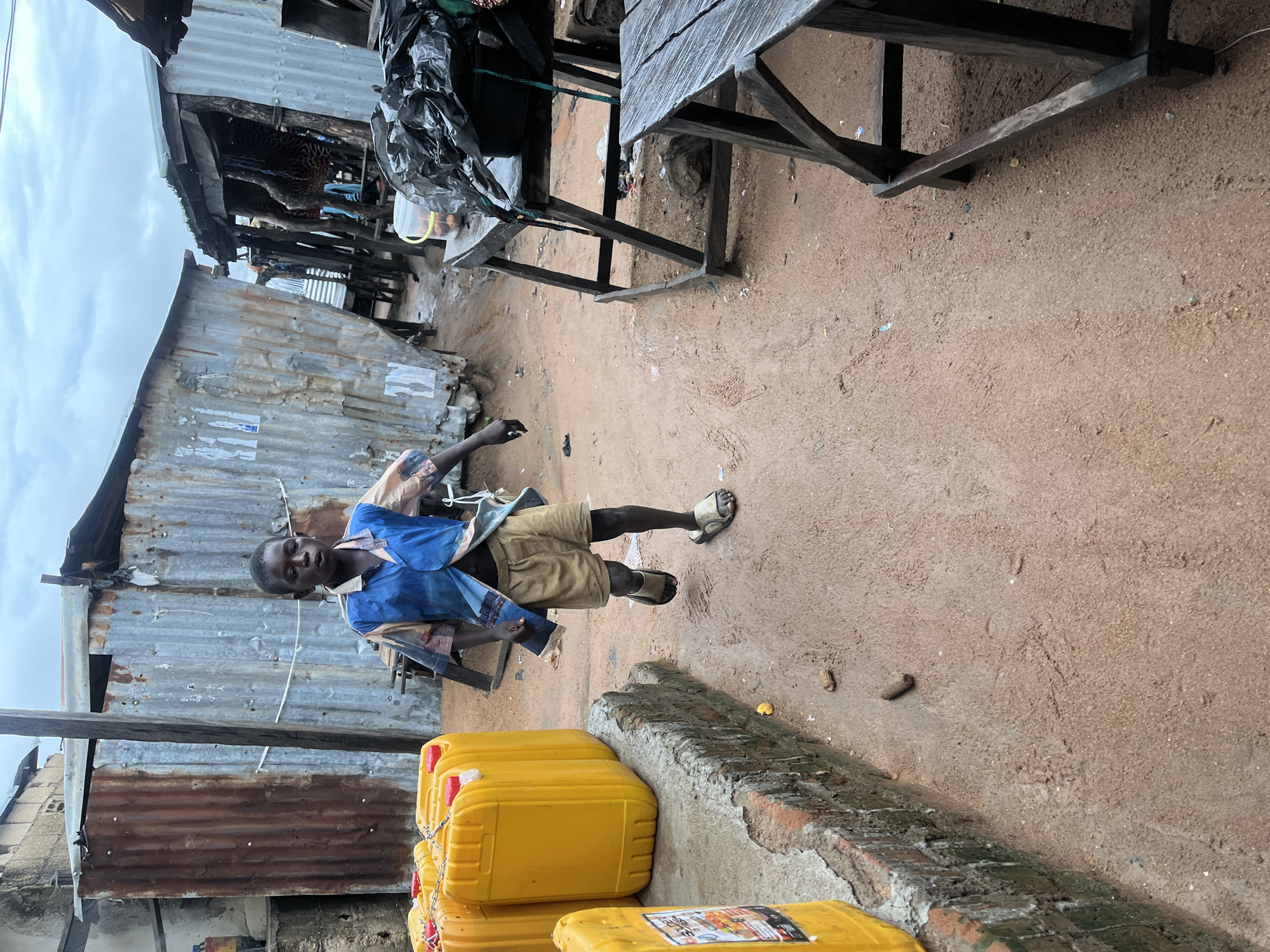 A young boy runs through a dirt path in Jalingo, Nigeria. He is wearing a blue and patterned shirt and khaki shorts, with sandals on his feet. The boy is running past several yellow jerrycans stacked on a low wall, and corrugated metal shacks line the background. The sky is overcast.