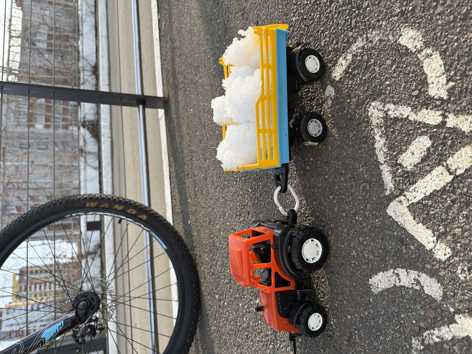 An outdoor, eye-level scene in Izhevsk, Russia, depicts a miniature plastic toy tractor pulling a trailer filled with snow on an asphalt surface. The toy tractor is predominantly orange with black chassis and white wheel hubs, hitched by a white plastic connector to a yellow and light blue toy trailer. The trailer's bed contains an irregular mound of white, granular material consistent with snow or crushed ice.
In the upper-left background, a portion of a bicycle's front wheel and fork is visible. The black tire bears "WANDA" branding and a blue stripe, while the black fork features a "LOKALN" decal and disc brake components. Behind the toys and bicycle, a dark grey metal railing, composed of vertical posts and horizontal wires or bars, spans the midground. Beyond this railing, an urban environment unfolds under a clear sky. Multi-story residential buildings with visible windows and balconies are present, intermingled with leafless trees, suggesting a winter or early spring season. The asphalt surface in the foreground displays partial white painted line markings. Strong shadows indicate bright daylight conditions. No people are visible.