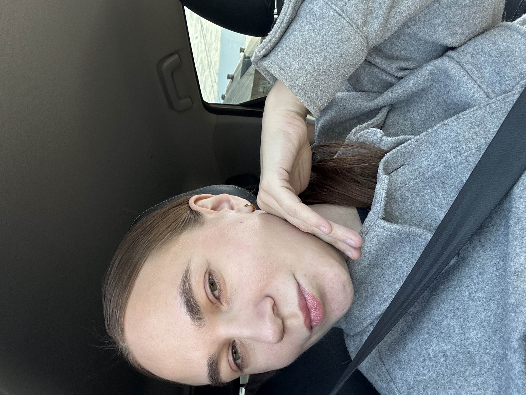 A young woman is seated in a car, wearing a gray coat and seatbelt. She is holding her face with one hand and looking towards the camera. The interior of the car is visible, including a grab handle. The location is not discernible from this image.
