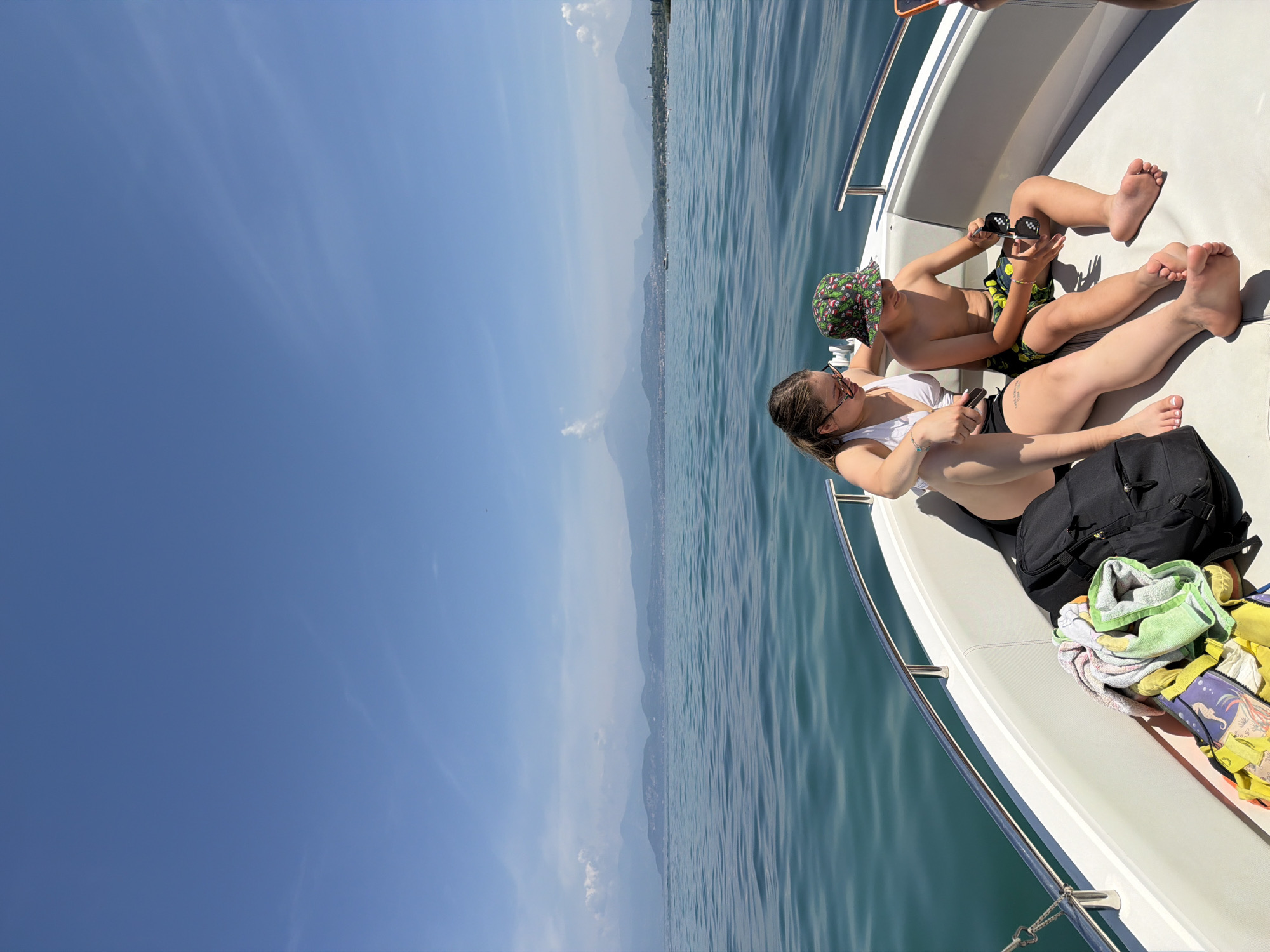 The image captures a sunny day on a boat in the waters near Peschiera del Garda, Italy. Two people, a woman and a boy, are seated on the front of the boat, facing forward. The woman, wearing a white bikini top and black shorts, has sunglasses on her head and her left arm resting on her lap. The boy, wearing patterned swim trunks and a bucket hat, holds a pair of sunglasses in his hands. In front of them, a black backpack and a pile of towels are visible. The boat is on a body of water with a clear blue sky overhead. In the distance, a chain of mountains lines the horizon, with some buildings and vegetation visible along the shore. The water is calm and reflects the blue of the sky.