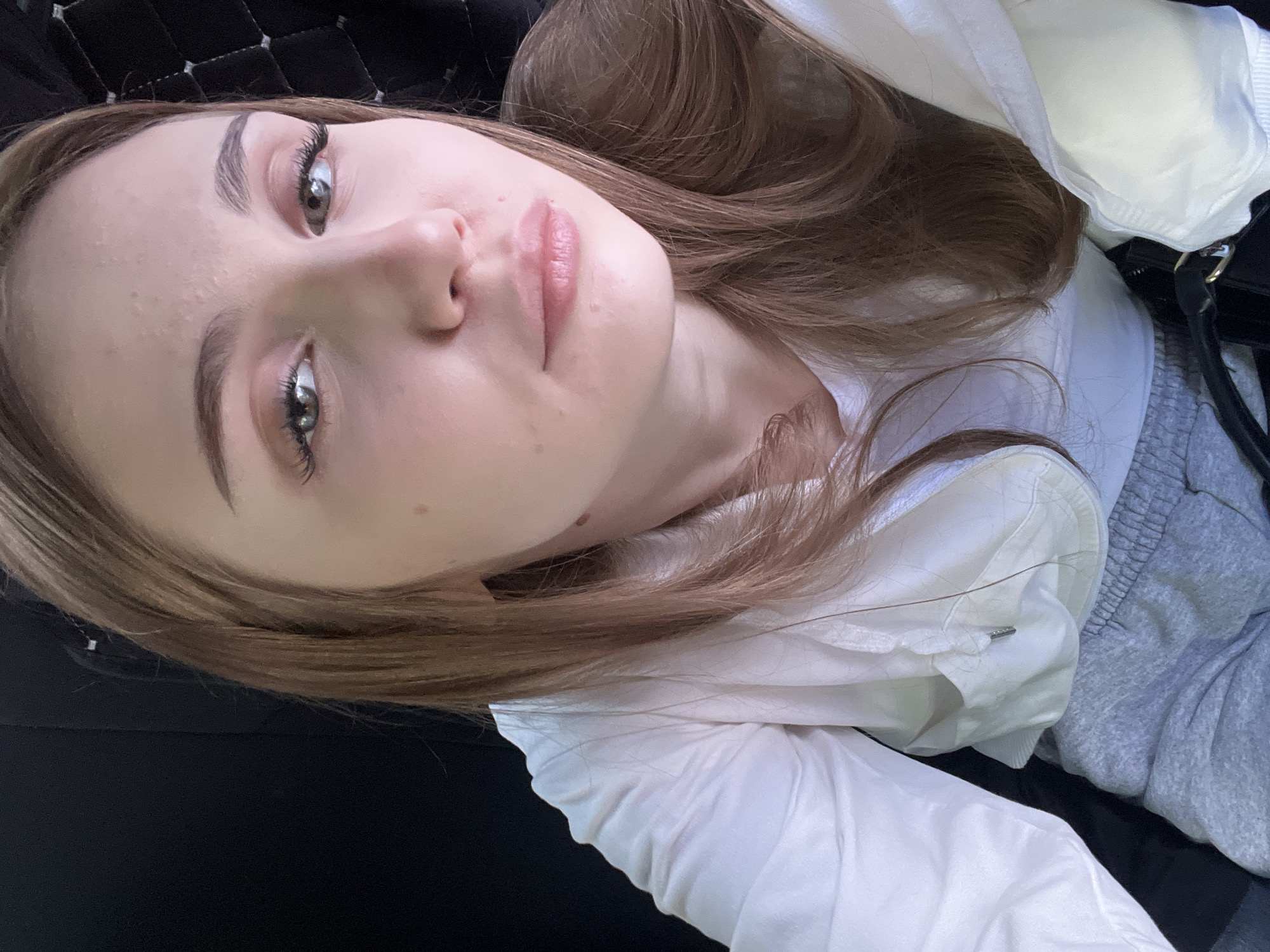 A close-up, high-angle selfie of a young Caucasian female with light brown hair and light green eyes. She is looking directly at the camera. She is wearing a white hooded jacket over a light gray t-shirt and gray sweatpants. Her hair is long and wavy, framing her face. She is wearing subtle makeup, including mascara and lip gloss. The background is dark and indistinct, suggesting an indoor setting, possibly a car interior, with some quilted texture visible on the right. There are no visible landmarks or environmental cues to indicate the specific location within the city of Tolbazy, Russia.