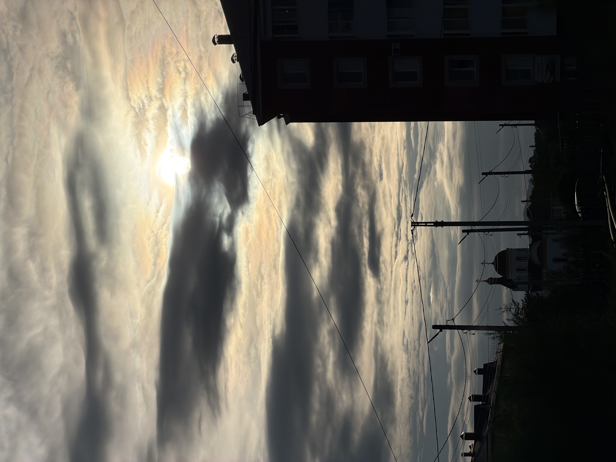 This image, captured in the city of Birobidzhan, Russia, presents an urban or suburban landscape under an overcast sky with high contrast. The sun is partially visible through a break in a dense cloud bank in the upper-middle section, casting a bright, slightly iridescent light that illuminates surrounding clouds with warm yellow and orange hues. Dark, layered clouds extend across the entirety of the sky, creating a somber yet luminous atmosphere.
In the right foreground, a multi-story building, likely residential, is sharply silhouetted, revealing a dark facade and a gabled roof. The roof features at least two distinct chimneys with rounded caps and a metal railing structure along its edge. Several rectangular windows are visible on the building's facade, appearing lighter than the surrounding structure due to internal lighting or reflection.
Multiple utility poles, both wooden and metal, are vertically positioned across the middle and lower-middle sections of the image. Numerous power lines stretch horizontally and diagonally between these poles, and one prominent dark power line extends from the upper-left corner diagonally downwards towards the right.
In the left-center midground, an Orthodox church is identifiable by its distinctive architecture, including a prominent golden onion dome topped with a cross, and at least one smaller, similarly designed dome. The church's structure appears predominantly as a silhouette, though some lighter tones suggest its underlying color. The immediate foreground consists of dark, indistinct masses of foliage and ground cover, obscuring the lower portions of the scene and the bases of the utility poles. No people are discernible in the image.