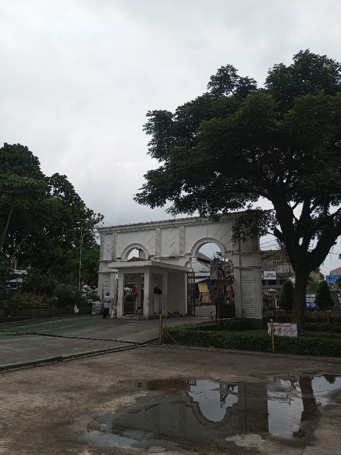 The image depicts an outdoor scene in Daringbadi, India, under an overcast sky. A prominent white neoclassical-style entrance structure stands centrally, featuring a covered central archway flanked by two open archways. The structure's facade is detailed with vertical fluting and arched recesses. To the left and right, large trees with dense green foliage partially frame the structure. The foreground consists of a gray paved area, where a reflective puddle shows a distorted reflection of the white structure. A single individual, dressed in light-colored clothing, is visible near the central archway of the structure, facing away from the viewer. Through the right archway, a building is partially visible, displaying a sign with "TOKO YOE YOE" written on it. To the right of the paved area, partially obscured by green shrubbery, a small white sign on a golden pole reads text in Indonesian, including "PERHATIAN DILARANG MEROKOK" (Attention No Smoking). The overall environment appears to be an urban or semi-urban setting.