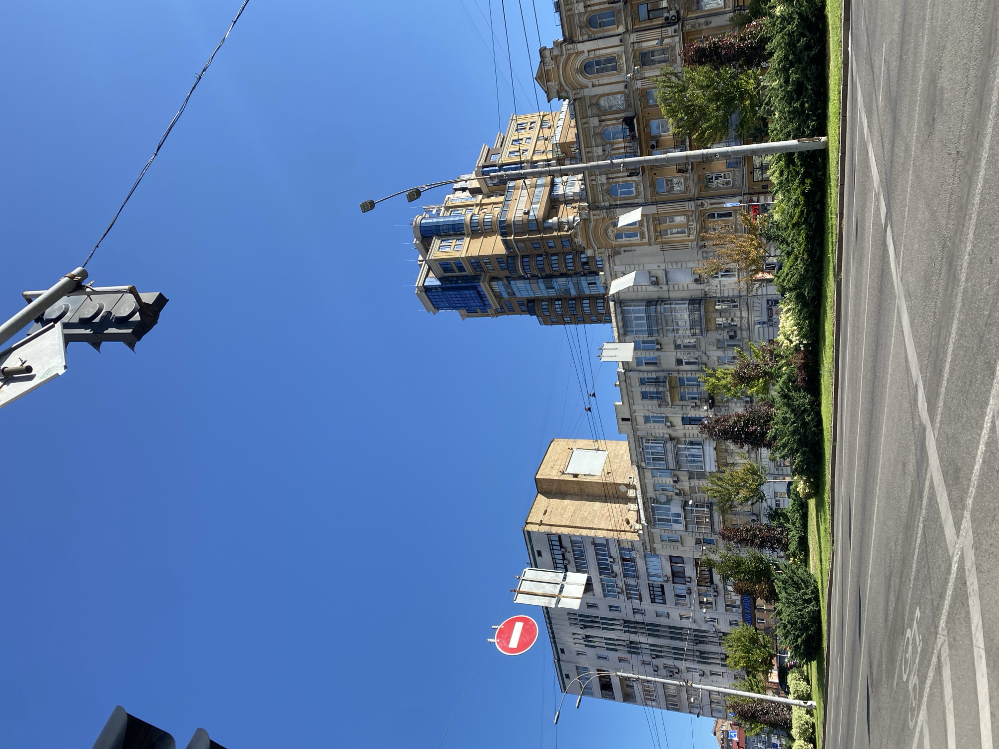 The image shows a street scene with buildings and a clear blue sky. In the foreground, a road with lane markings is visible. A lush green median separates the road from the sidewalk. Several trees and shrubs are planted along the median.
Behind the median, a row of apartment buildings stands. The buildings vary in architectural style and height. The leftmost building is a tall, modern apartment block with a facade of white and gray panels, featuring numerous windows and balconies. To its right, a mid-rise building with a textured yellow facade is visible. Further to the right, a series of older, ornate buildings with yellow and cream facades are present. These buildings feature detailed architectural elements like arches and balconies. A particularly tall, modern building with a glass facade is situated behind some of the older structures, dominating the skyline.
Above the buildings, a traffic light hangs over the road, and electrical wires are strung across the scene. A circular red and white traffic sign, indicating "no entry" or "do not enter," is suspended from a wire. A lamppost is also visible. The overall impression is a sunny day in an urban environment.
