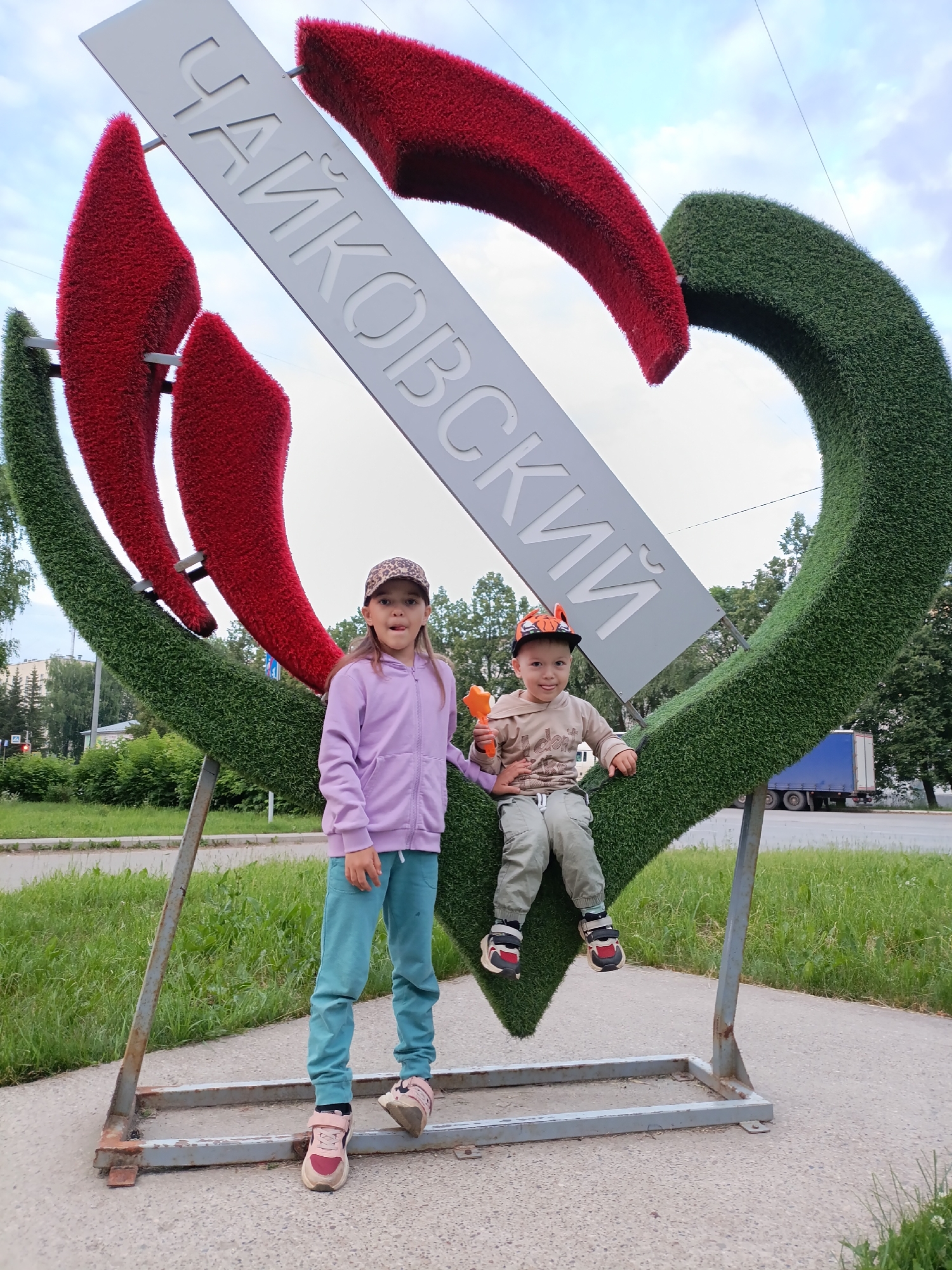 A girl and a boy pose in front of a large, heart-shaped sculpture adorned with green and red artificial grass, spelling out "ЧАЙКОВСКИЙ" (Chaikovsky) in Cyrillic letters. The girl, wearing a purple hoodie and turquoise pants, stands to the left. The boy, in a beige hoodie and light green pants, sits on the sculpture to the right, holding an orange toy. The sculpture appears to be a landmark in the city of Chaikovsky, Russia, situated outdoors with a street and trees visible in the background. The sky is overcast, suggesting cloudy weather.