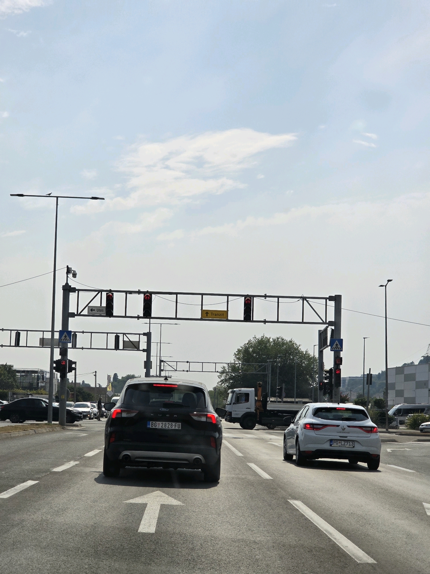 The image depicts a street scene in Podgorica, Montenegro, likely an intersection, under a bright, overcast sky with scattered clouds. In the foreground, two cars are visible on a multi-lane asphalt road marked with white dashed lines. The car closest to the viewer is a black SUV with its taillights illuminated. The license plate reads "BG 2828-FU". To its right, a white hatchback is moving away from the viewer. Its license plate reads "PG-LZ713". A large white arrow is painted on the road, indicating a forward direction.
Overhead, a traffic signal gantry spans the road, displaying red lights on multiple traffic signals. Signs on the gantry indicate directions: one with an arrow pointing left and the text "Ubli", and another with an arrow pointing up and the text "Tranzit". A pedestrian crossing sign with a blue background and a white silhouette of a person walking is also visible.
In the mid-ground, a white flatbed truck is stopped or moving slowly. Behind the truck, a large green tree is prominent. In the background, buildings are visible, including a multi-story structure with a checkered facade on the right. Other cars and streetlights are also present. A small bird is perched on a streetlight pole on the left side of the image. The overall impression is of a typical urban road environment during daytime.