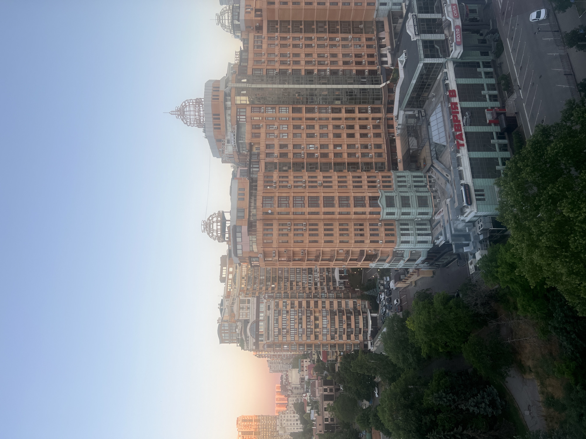 The image is an elevated, medium shot of a city skyline at dusk, featuring a cluster of tall, modern residential buildings in Odesa, Ukraine. The buildings are predominantly made of light-colored brick with numerous windows and air conditioning units visible. Several buildings feature distinctive dome-like structures on their rooftops.
In the foreground, a large building with a green and glass facade has signage indicating "ТАВРІЯ В" (Tavria V) and "ФІТНЕС-ПРОСТІР" (Fitness-Prostir), suggesting it's a commercial complex housing a supermarket and a fitness center. The parking lot in front of this complex is mostly empty, with a few cars scattered around. Adjacent to the commercial building, there's a street with parked cars and trees lining the sidewalks.
The background shows more residential buildings, some taller and more uniformly built, with a soft, warm glow from the setting sun illuminating the sky and the distant structures. The sky transitions from a pale blue at the top to a soft pink and orange hue near the horizon, indicative of twilight. Lush green trees are visible between the buildings and in the foreground, suggesting a well-maintained urban environment with green spaces. The overall impression is one of a modern, somewhat dense urban area in Odesa.