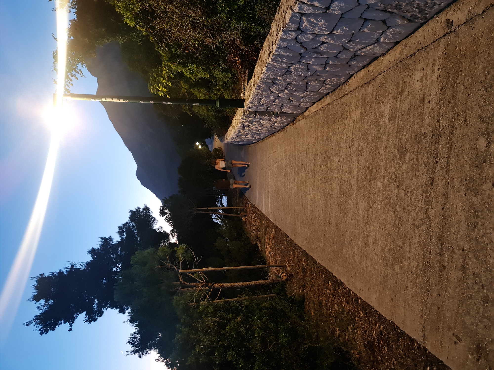 The image shows a paved walkway in the city of Brist, Croatia, at dusk. Two individuals, appearing to be women, are walking away from the camera on the walkway. The walkway is flanked on the right by a stone wall, and on the left by trees and foliage. A lamppost is visible on the right side of the walkway, casting light. In the background, mountains are visible, and the sky is a deep blue, with a thin white cloud formation streaking across it. The overall scene suggests a serene evening stroll in a coastal town.