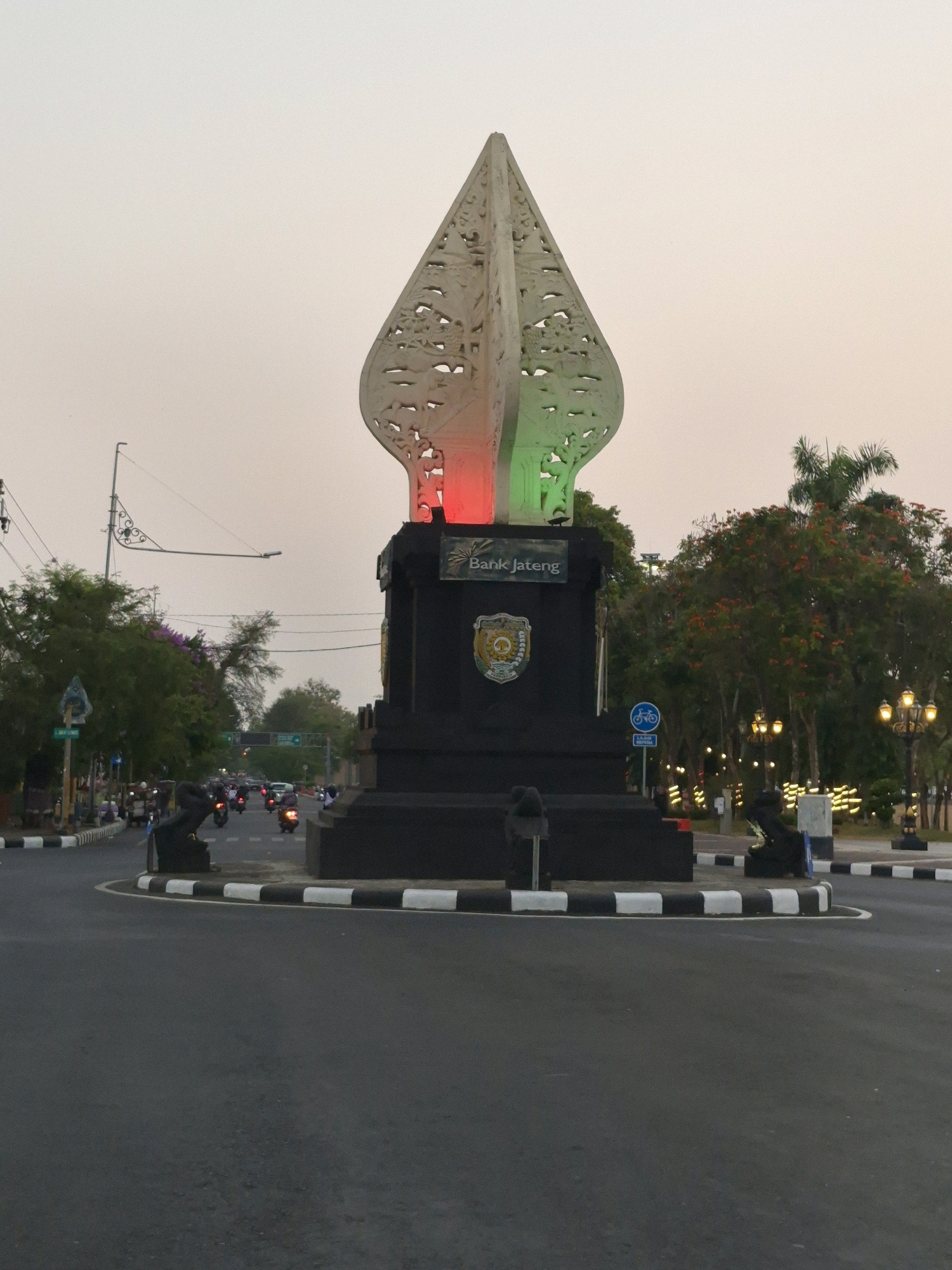 The image captures a view of a monument in the city of Purworejo, Indonesia, likely during the late afternoon or early evening. The monument is a dark, imposing structure topped with a stylized, white, leaf-shaped ornament illuminated with red and green lights. The text "Bank Jateng" is visible on the monument. The monument is situated in the center of a traffic circle with a distinct black and white checkered border. Several motorcycles are seen navigating the traffic circle and the surrounding roads, indicating active urban movement. Trees and streetlights line the background, suggesting a public space. The sky is overcast and light, consistent with dusk.