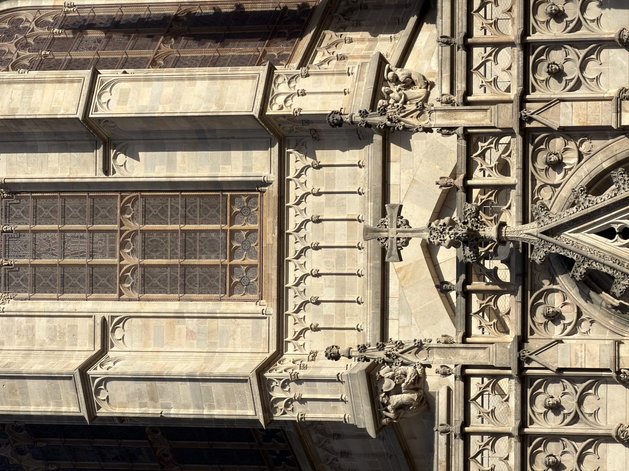 This is a close-up, overhead shot of the intricate Gothic architecture of a building in Barcelona, Spain. The sun is shining, casting dramatic shadows across the stone facade. The architecture features numerous carved elements, including gargoyles, ornate tracery in the windows, and decorative stone carvings of faces and figures. One prominent sculptural group appears to depict a scene with several figures. The stonework is a pale, weathered beige, contrasting with the darker, shadowed areas. The windows are tall and narrow, divided into multiple panes with leaded glass and intricate, lace-like stone surrounds. The overall impression is one of great age and detailed craftsmanship. The perspective is tilted, giving a dynamic and abstract feel to the composition. There is no visible text.